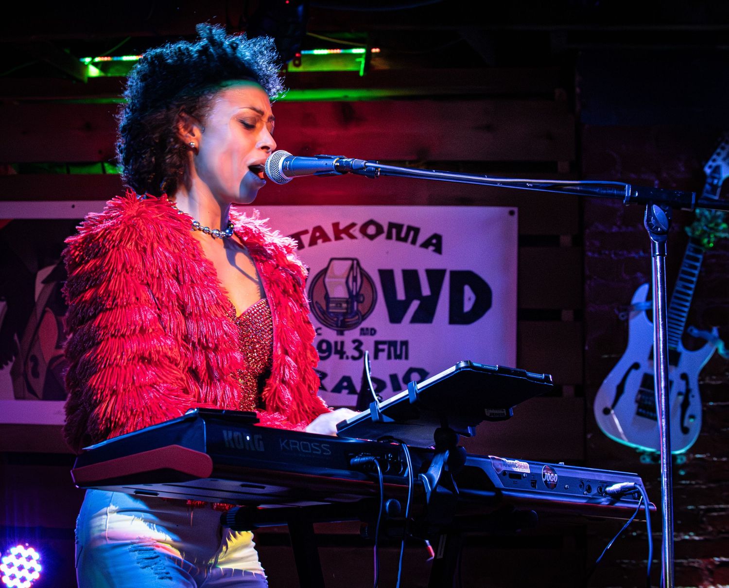 A musician performs on stage in a red feathered outfit while playing keyboard at a dimly lit music venue.