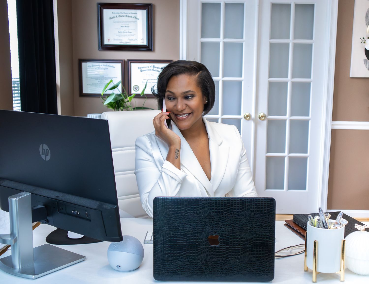 Professional woman in white blazer smiling while talking on phone at desk with HP monitor and laptop in modern office.