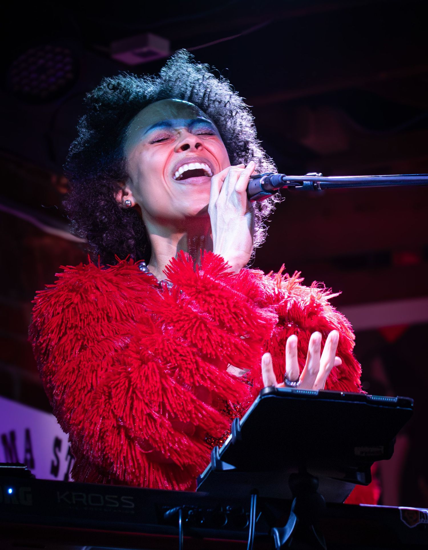 Performer in vibrant red feathered top sings passionately at microphone while performing at piano on dimly lit stage.