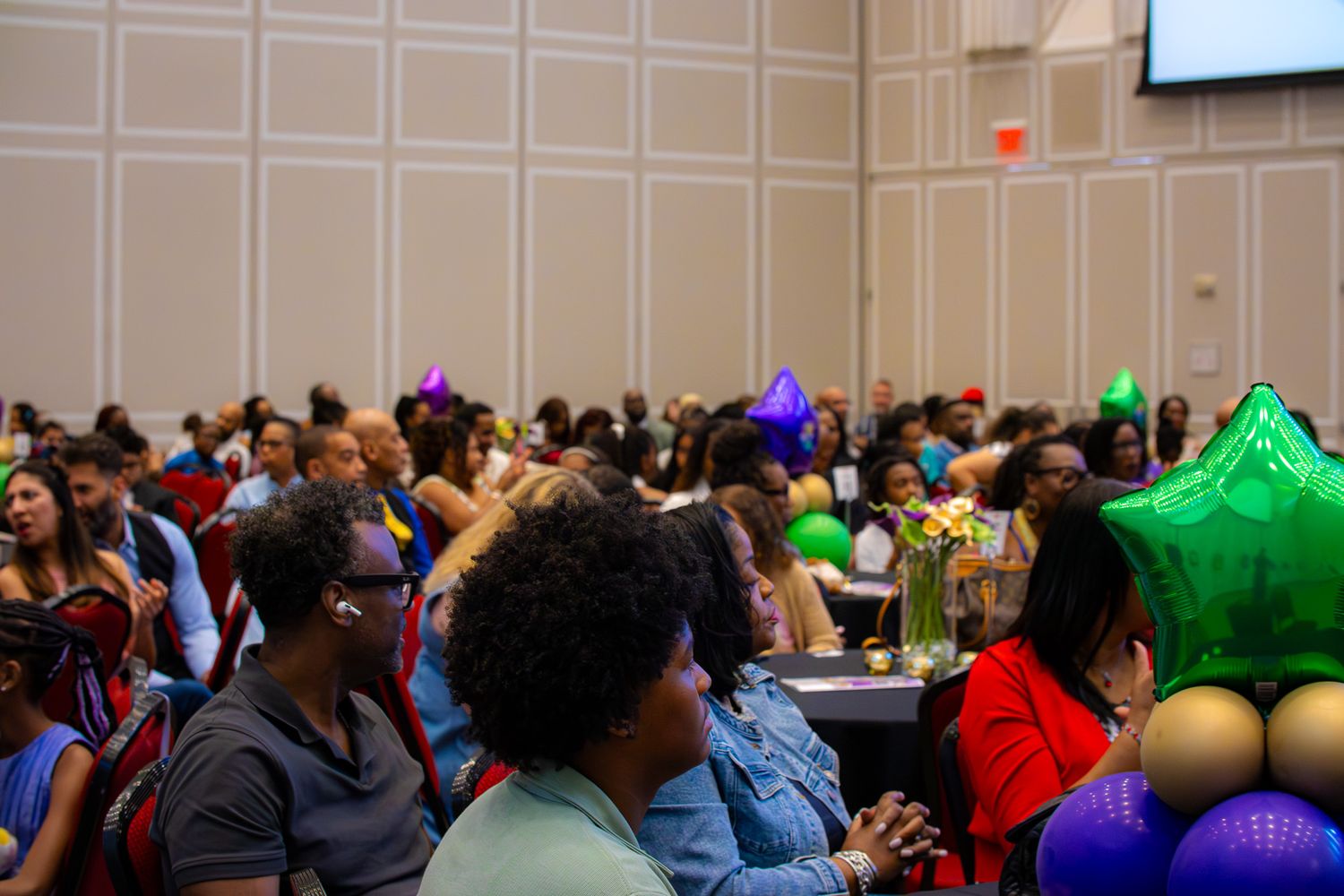 Large diverse group of people seated at tables in a ballroom event with colorful balloons and decorations.