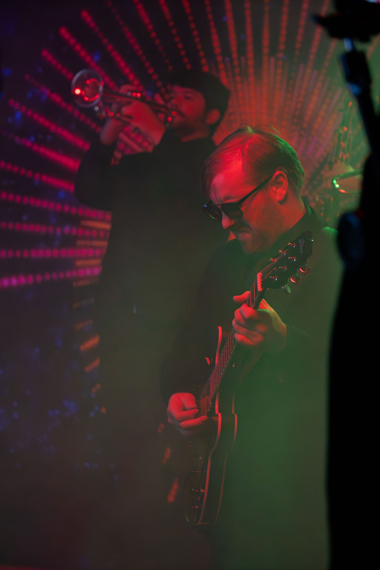 Musicians perform in dramatic red and green lighting with radial light pattern effect.