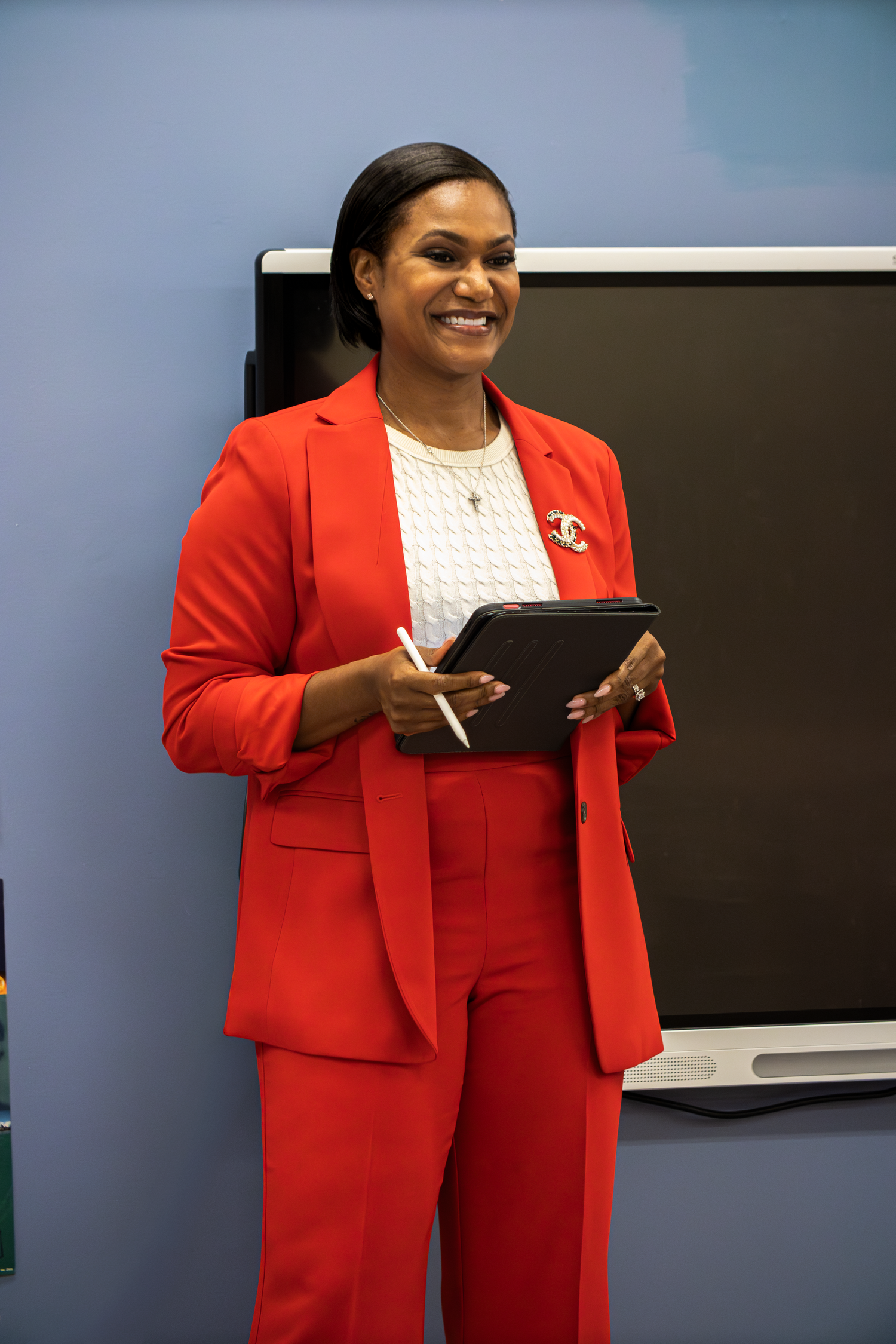 Professional woman in red pantsuit holds tablet and stylus while standing near smartboard.