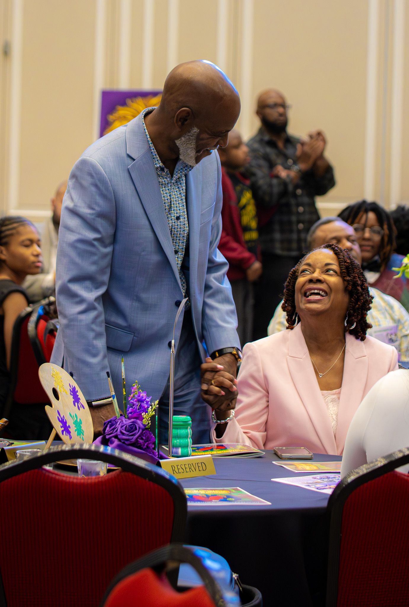 Man in blue blazer holds hands with smiling woman in pink blazer at a reserved banquet table during a celebration event.