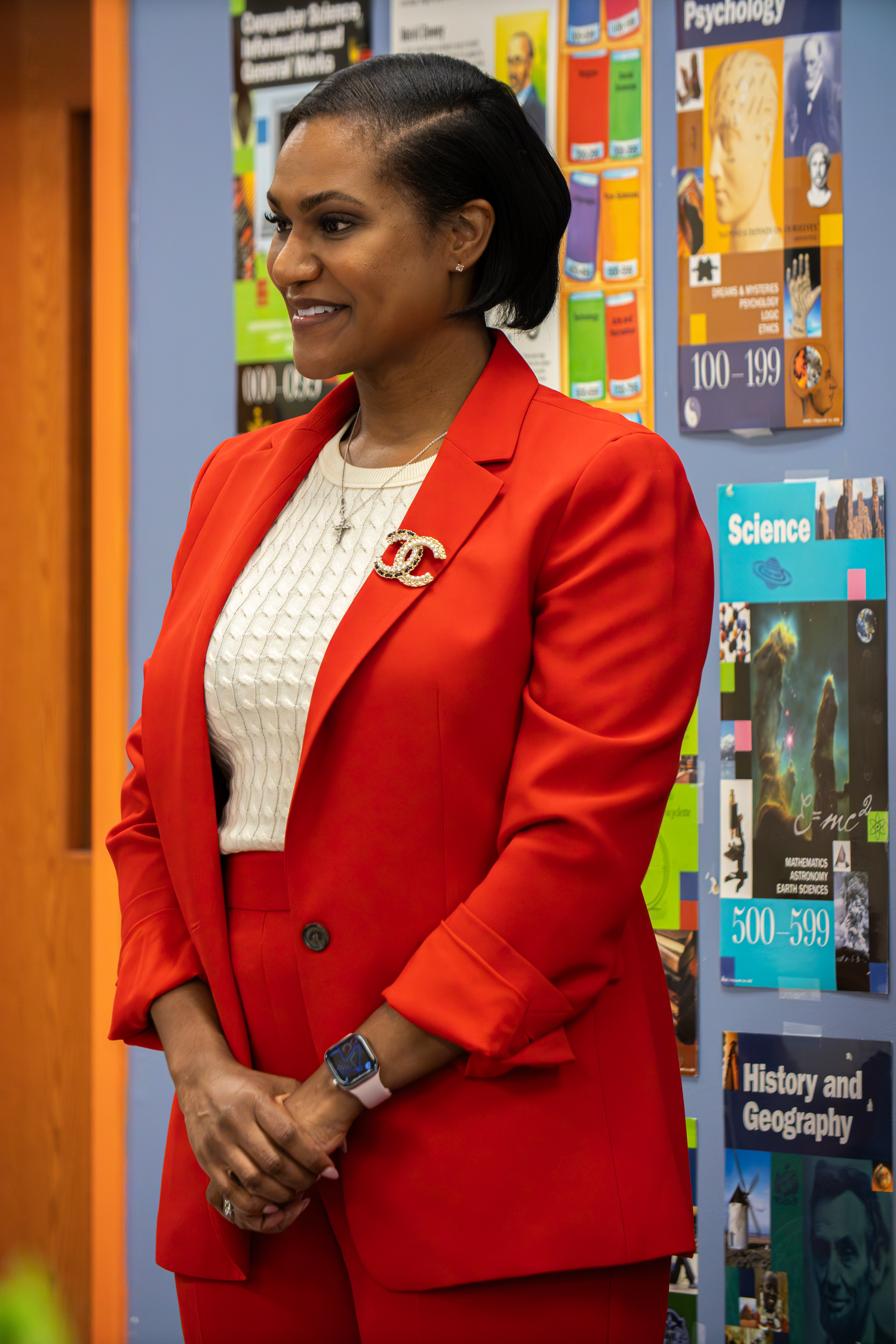 Professional woman in red blazer suit with Chanel brooch smiling in colorful library setting.