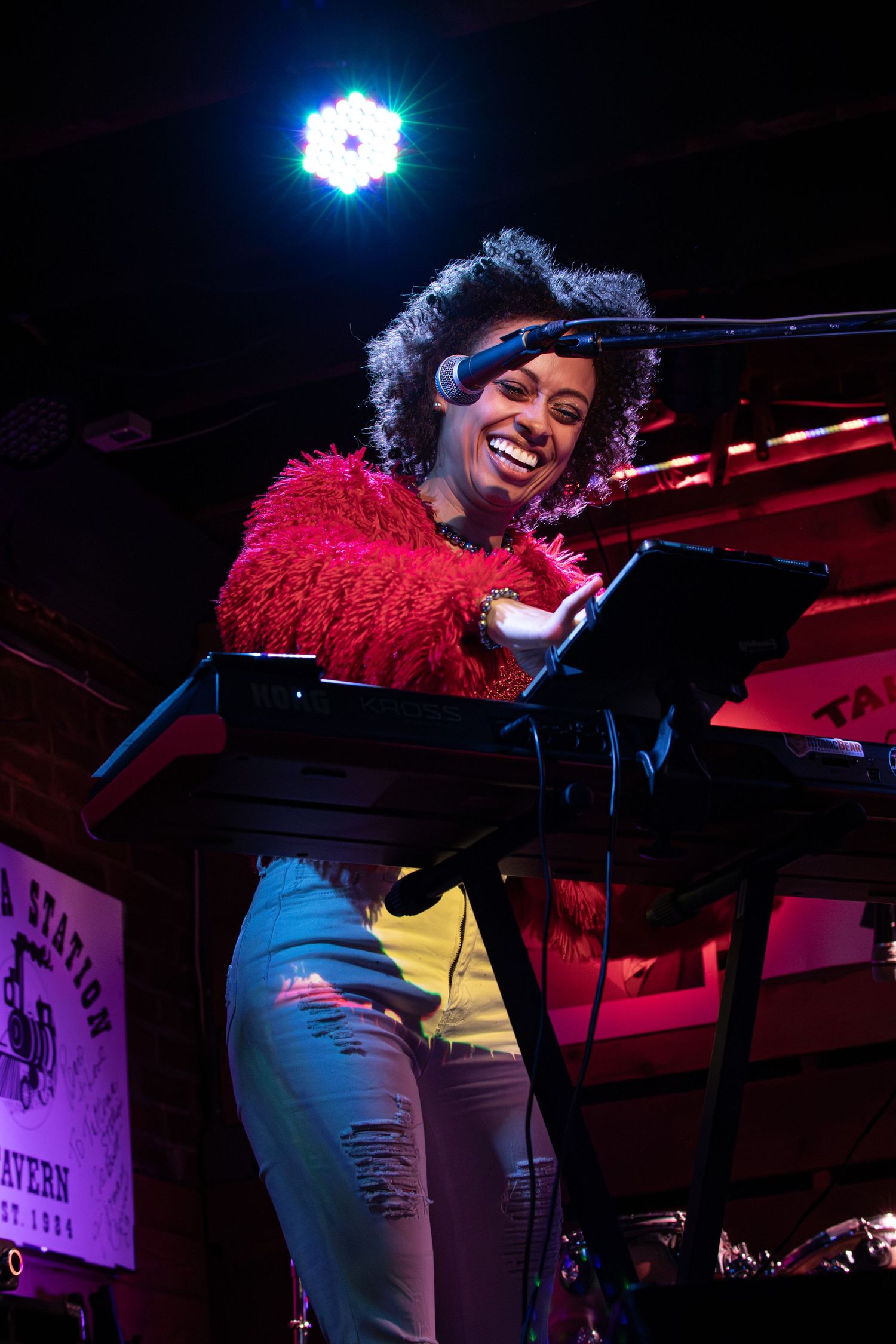 A performer in a vibrant red feathered top plays piano on stage under dramatic blue lighting.