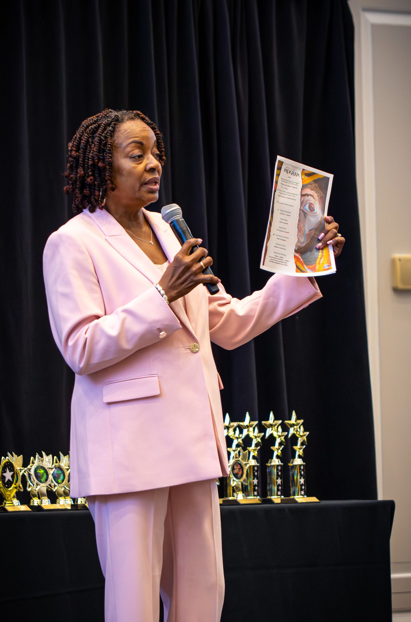 Woman in pink suit holding microphone and document at awards ceremony with trophies displayed in background.