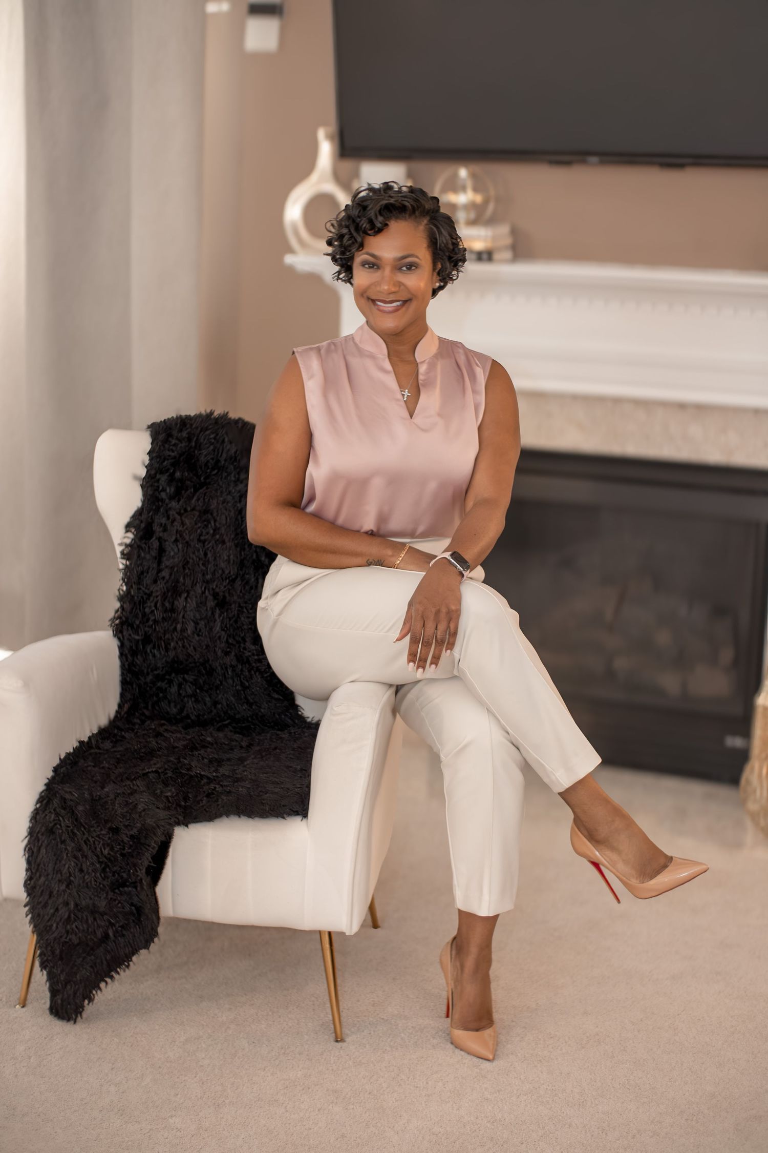A smiling woman in a pink blouse and white pants sits elegantly in a white chair beside a fireplace.
