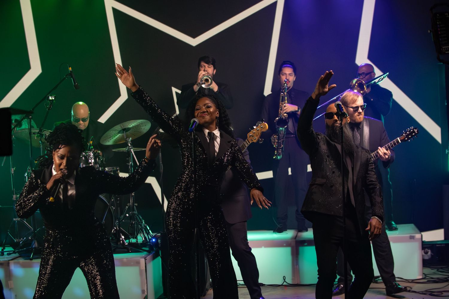 Band members perform on stage with geometric white lighting patterns in the background.