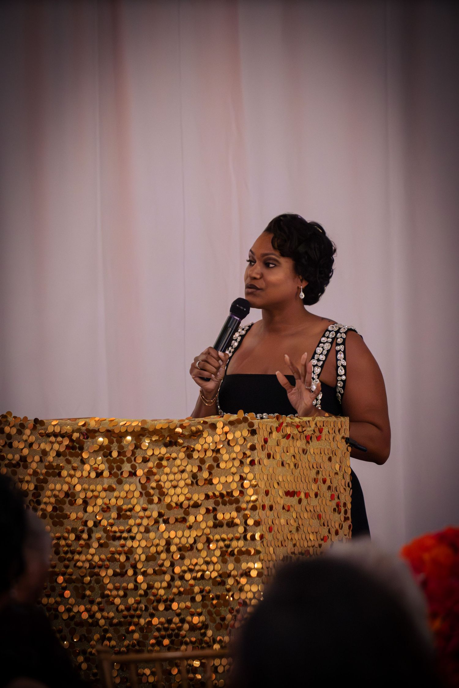 A performer in a black dress sings passionately on stage with a shimmering gold backdrop.
