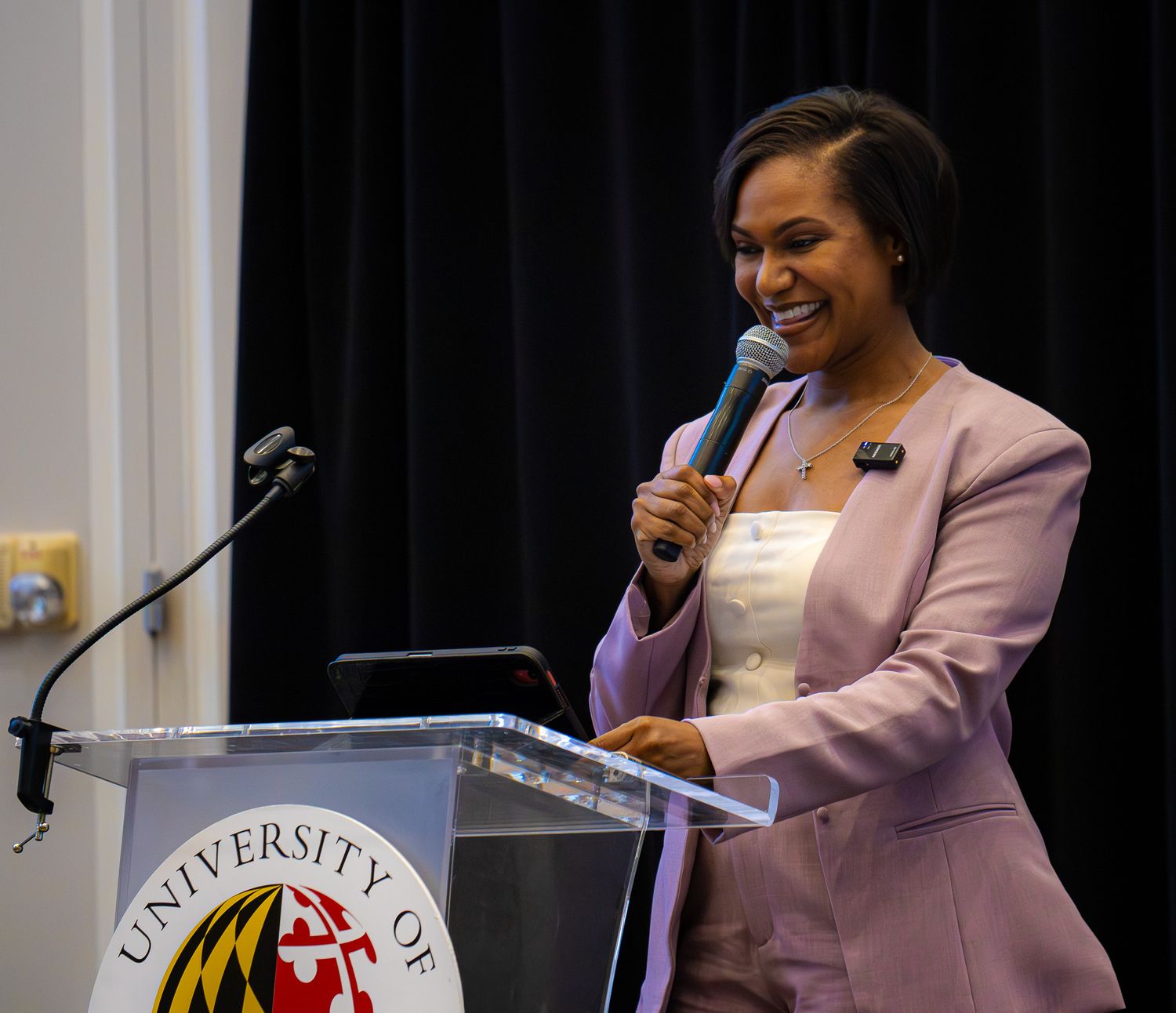 Woman in pink blazer speaking at University of Maryland podium, smiling while holding microphone at formal event.
