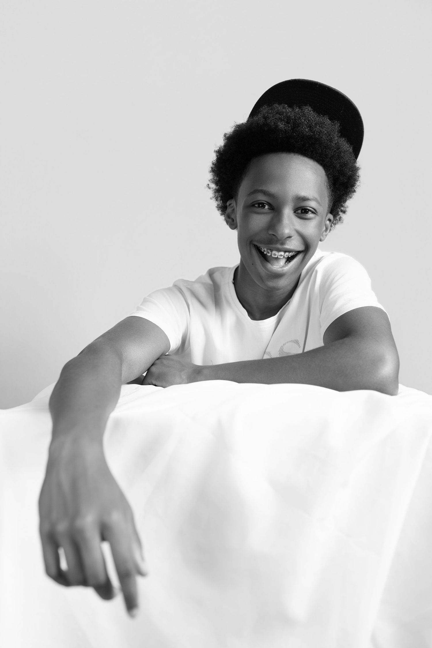 Smiling young Black teen with braces and a black hat leans over a white surface in a black and white portrait photo.