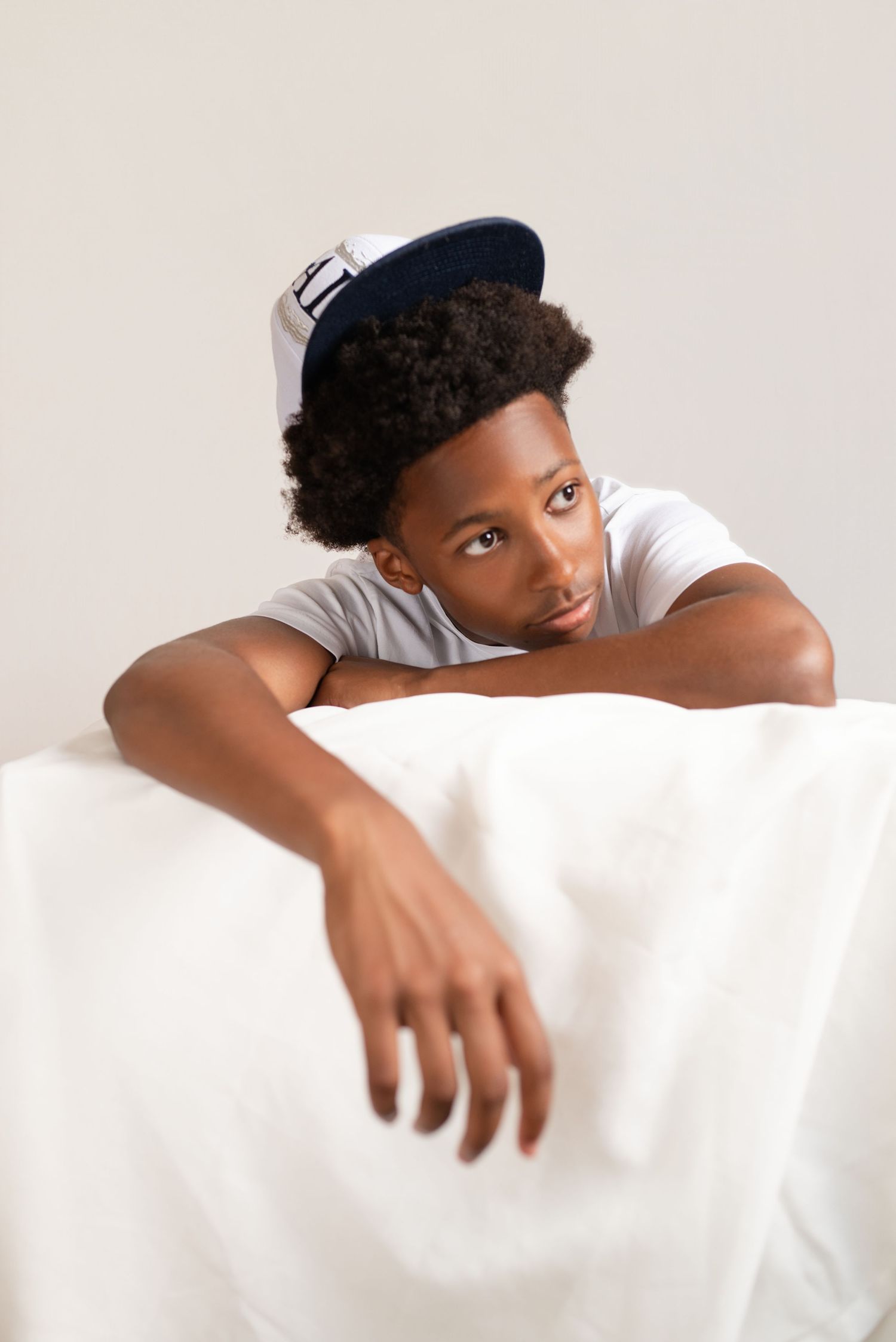 Young Black teen in white t-shirt and navy snapback cap leaning on white surface, looking thoughtful.