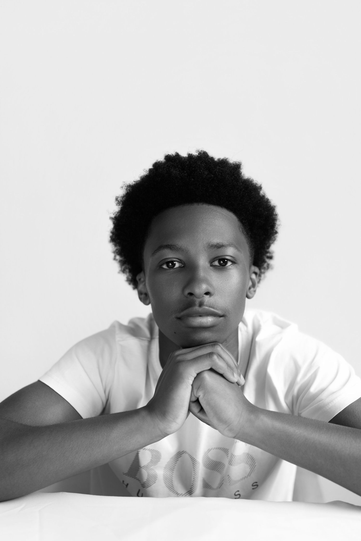 Black and white portrait of a young man with afro hair, hands clasped under chin, wearing a white BOSS t-shirt.