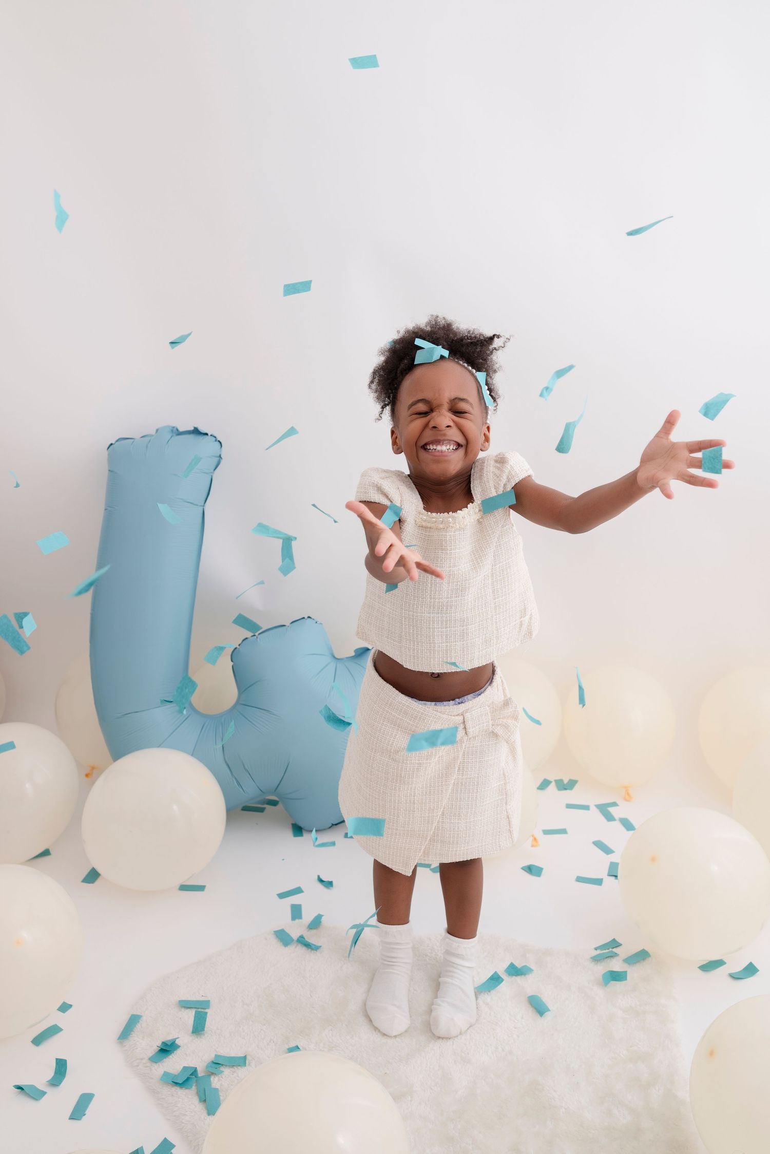 Child in white dress jumping joyfully with blue confetti falling in studio setting.