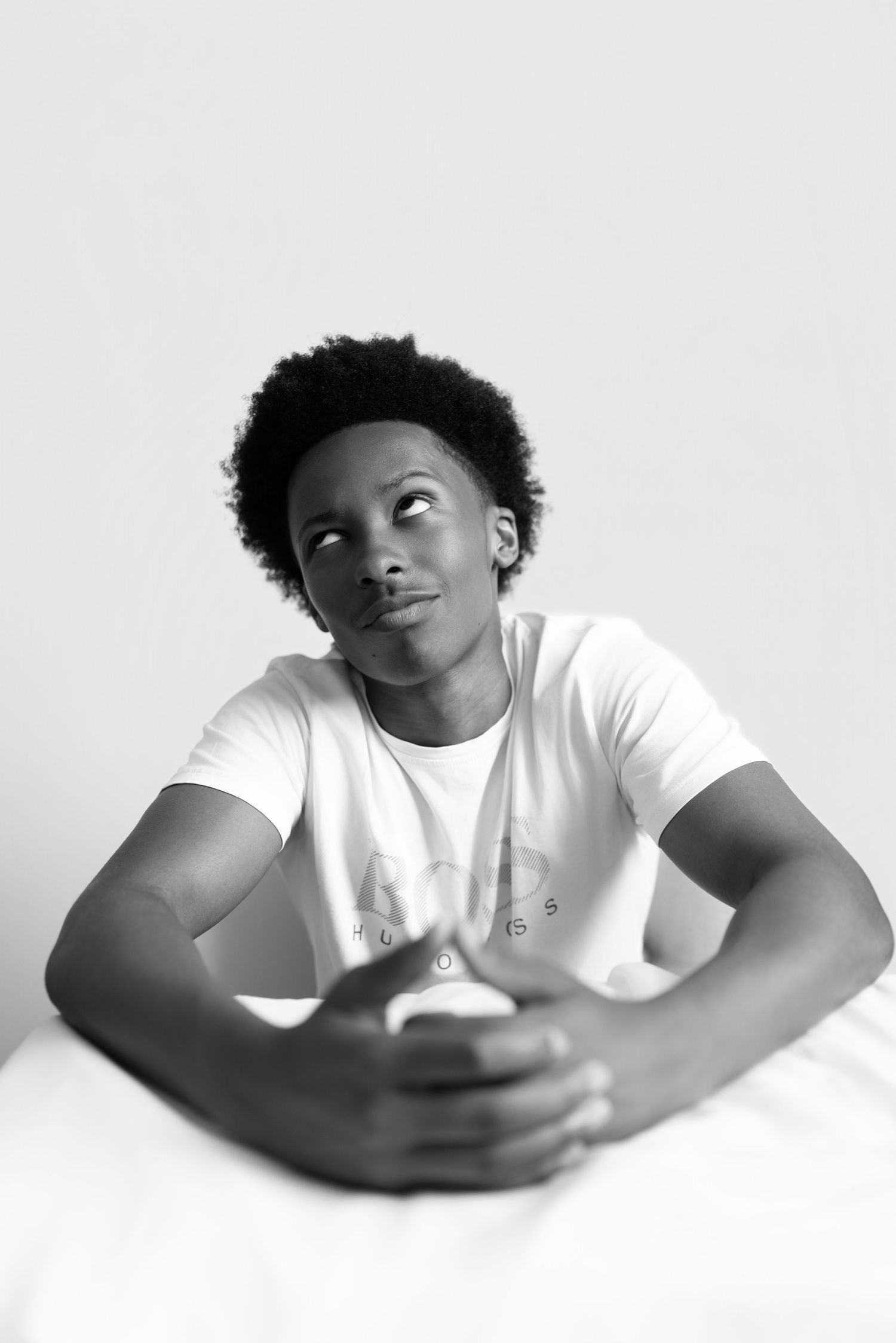 Black and white portrait of a young man in a white t-shirt sitting at a table, looking thoughtfully upward.