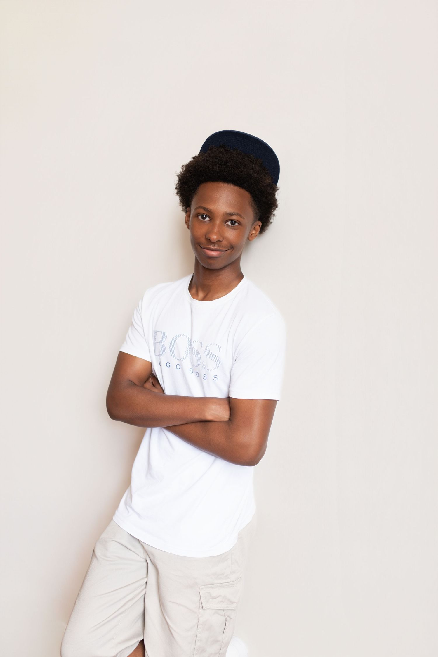Young Black teen in white BOSS t-shirt, navy cap, and khaki shorts, posing with arms crossed against a white wall.