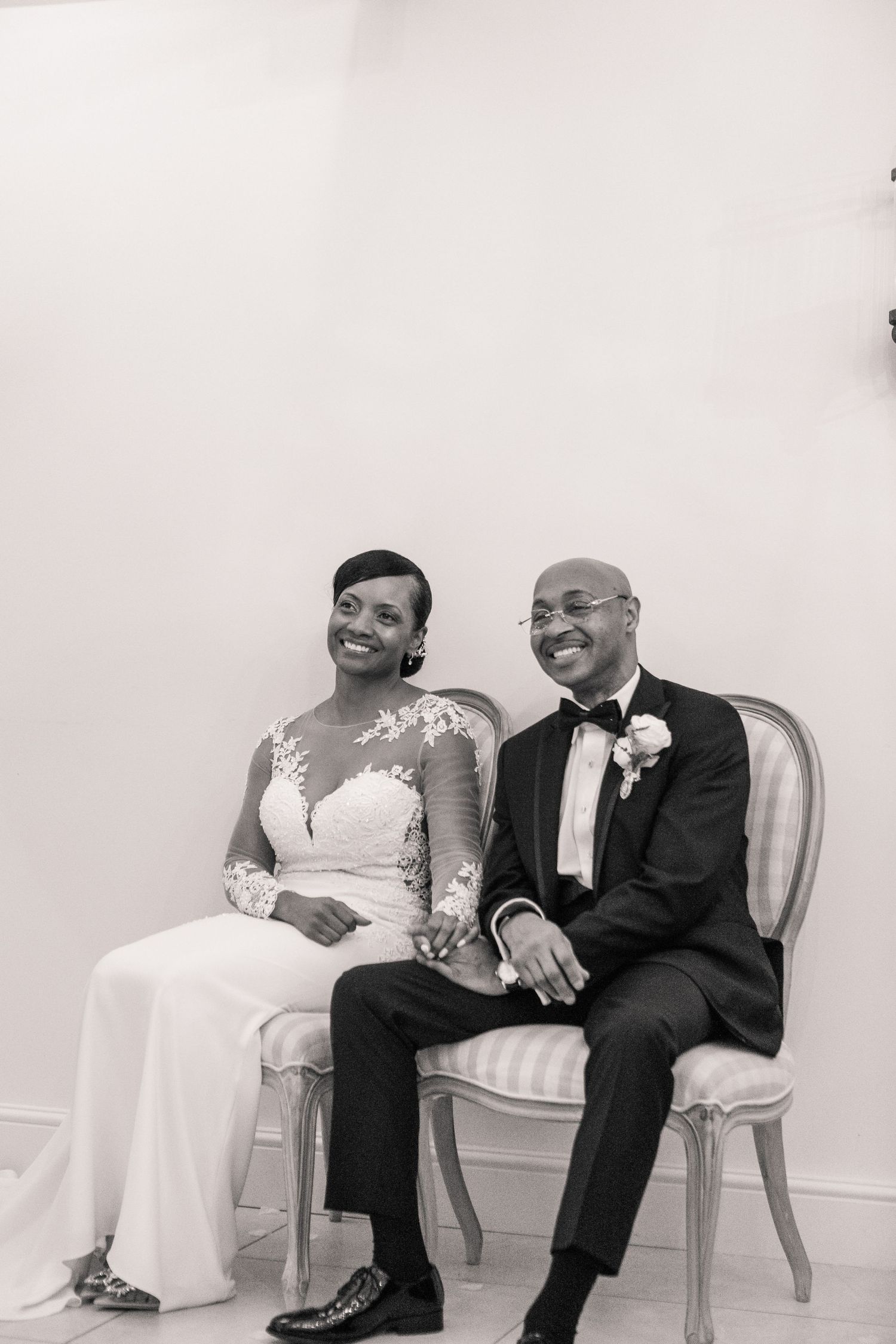 A couple shares a joyful moment while sitting together on elegant white chairs during what appears to be a wedding reception.