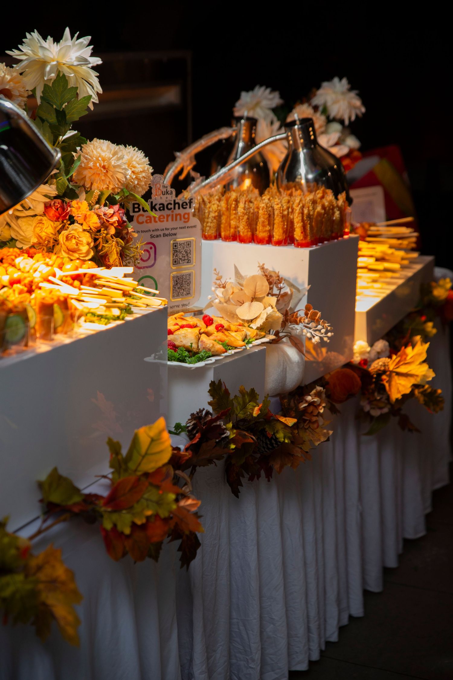 Elegant buffet table decorated with fall leaves and white linens displaying catered food items.