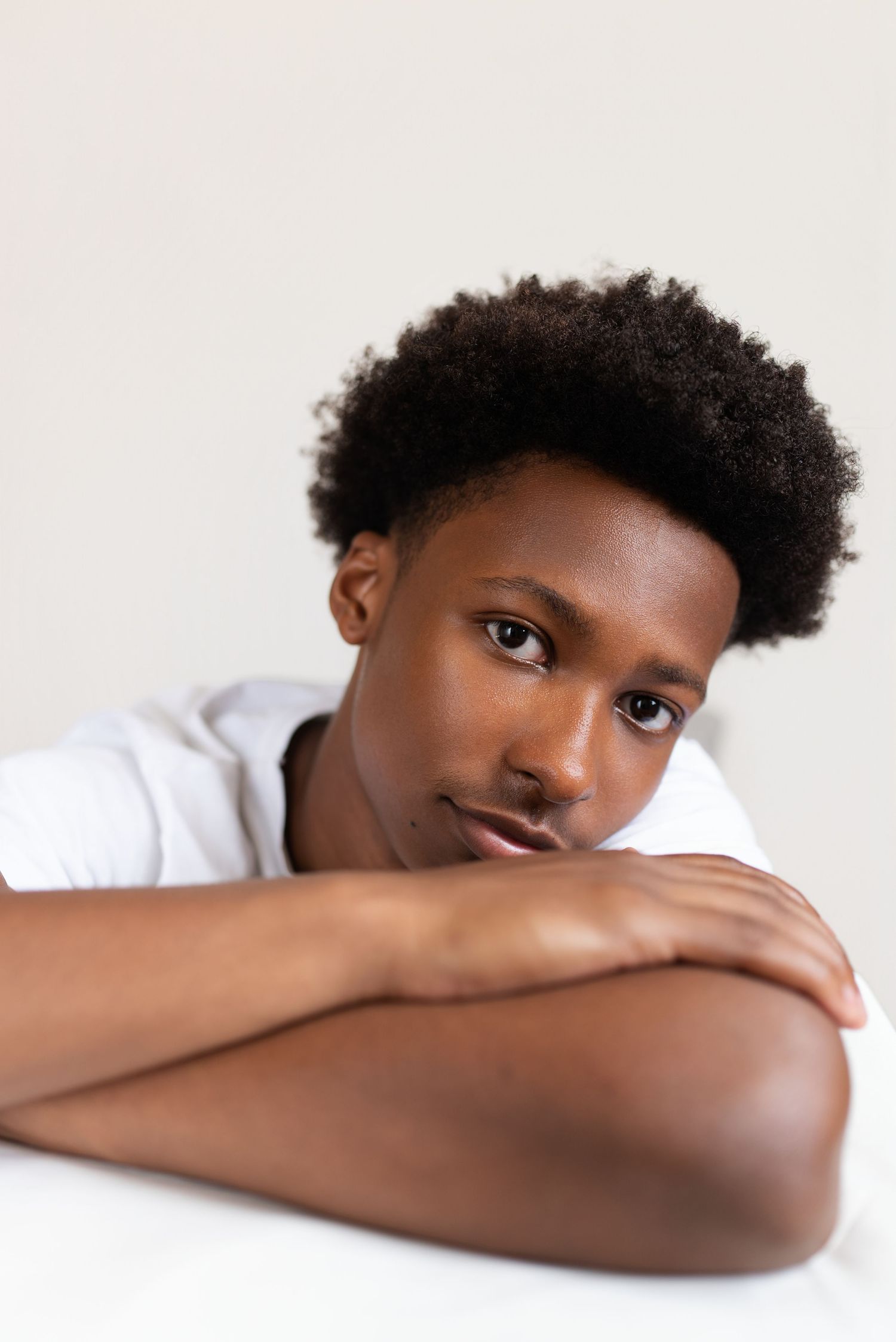 Young Black man with afro hairstyle resting arms on white surface, wearing white t-shirt, looking at camera.