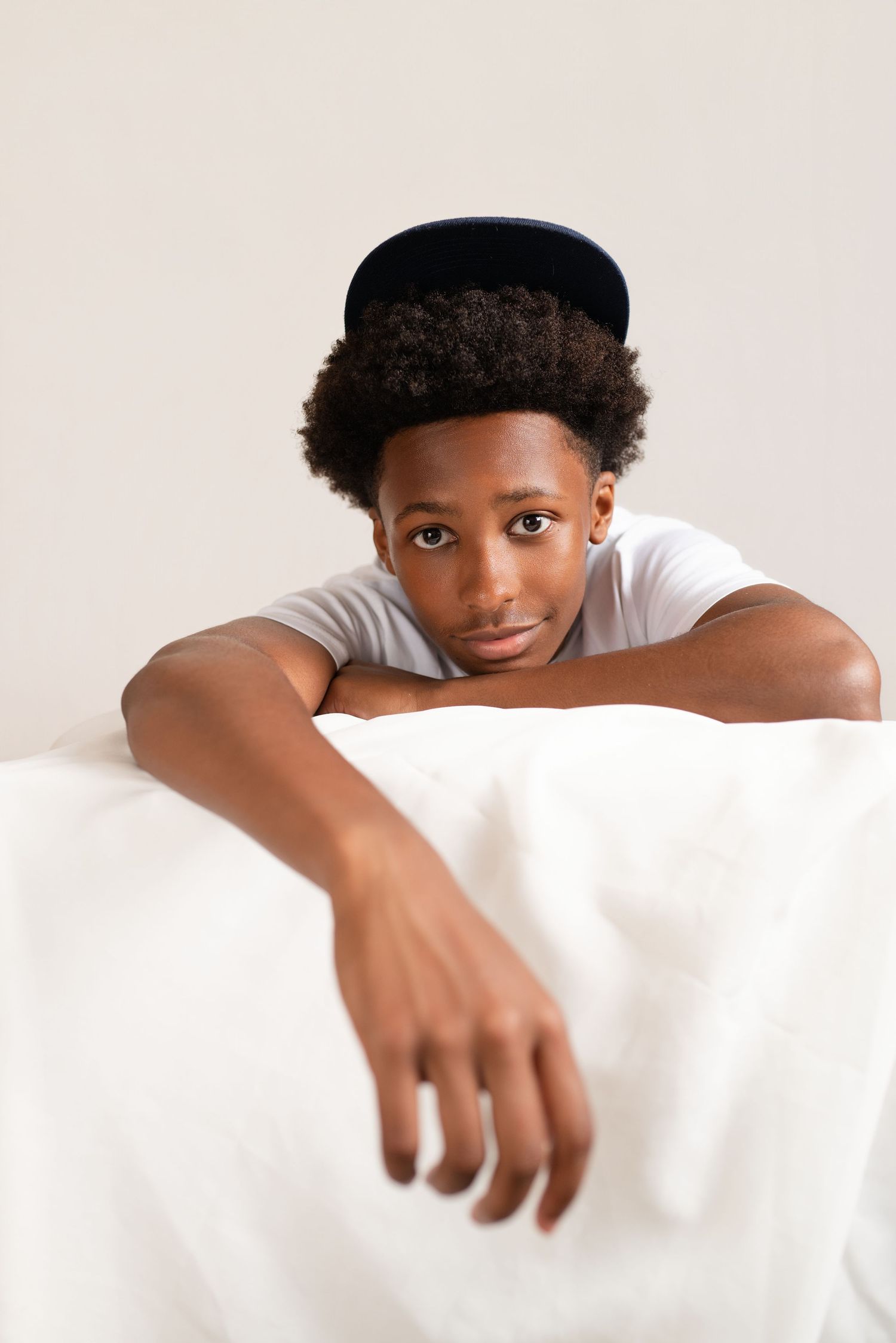 A young Black teen wearing a snapback hat and white t-shirt leans over a white surface, looking at the camera.