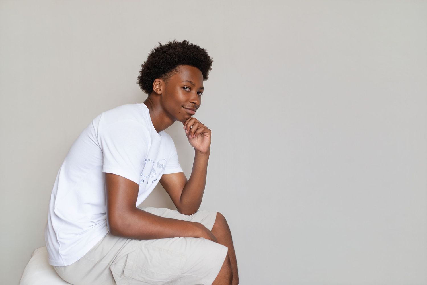 Young Black man in white t-shirt sitting thoughtfully with hand on chin against light gray background.