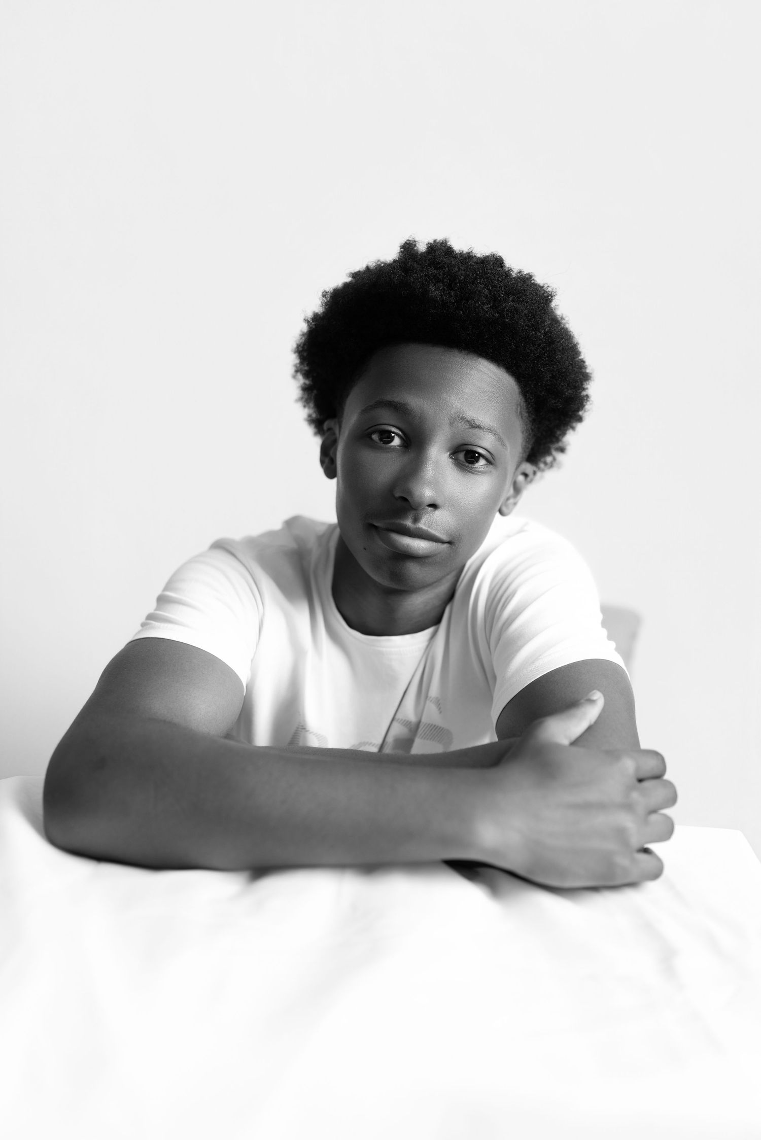 Black and white portrait of a young man with natural hair wearing a white t-shirt, resting arms on a surface.