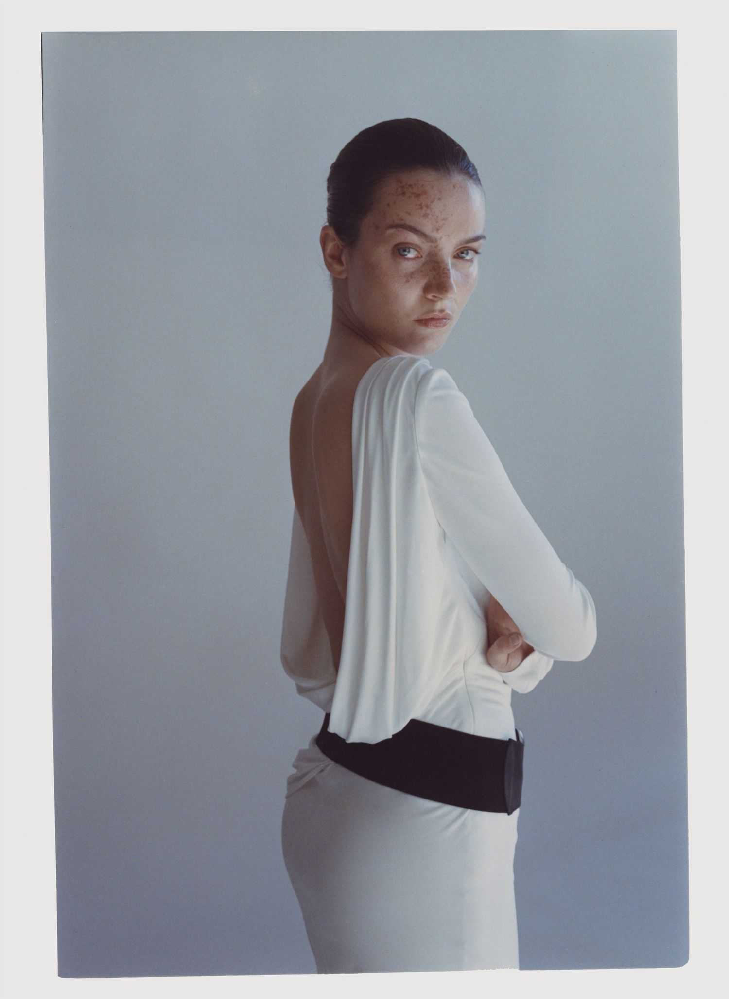 Film portrait of a model with freckles wearing a draped white dress and large black belt.