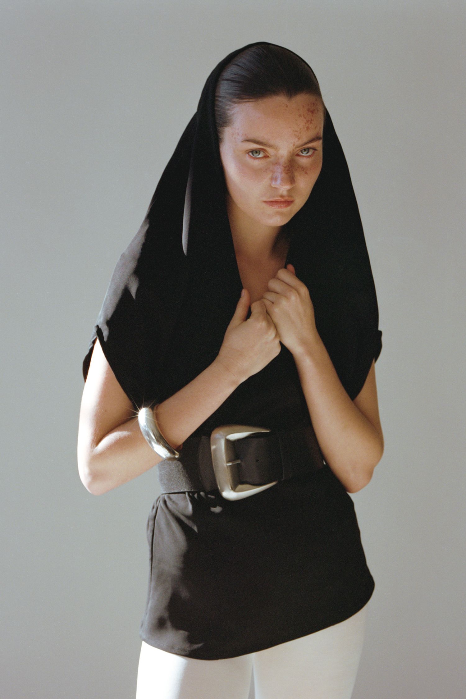 A model with freckles wearing a black hooded top and large black and brass belt.