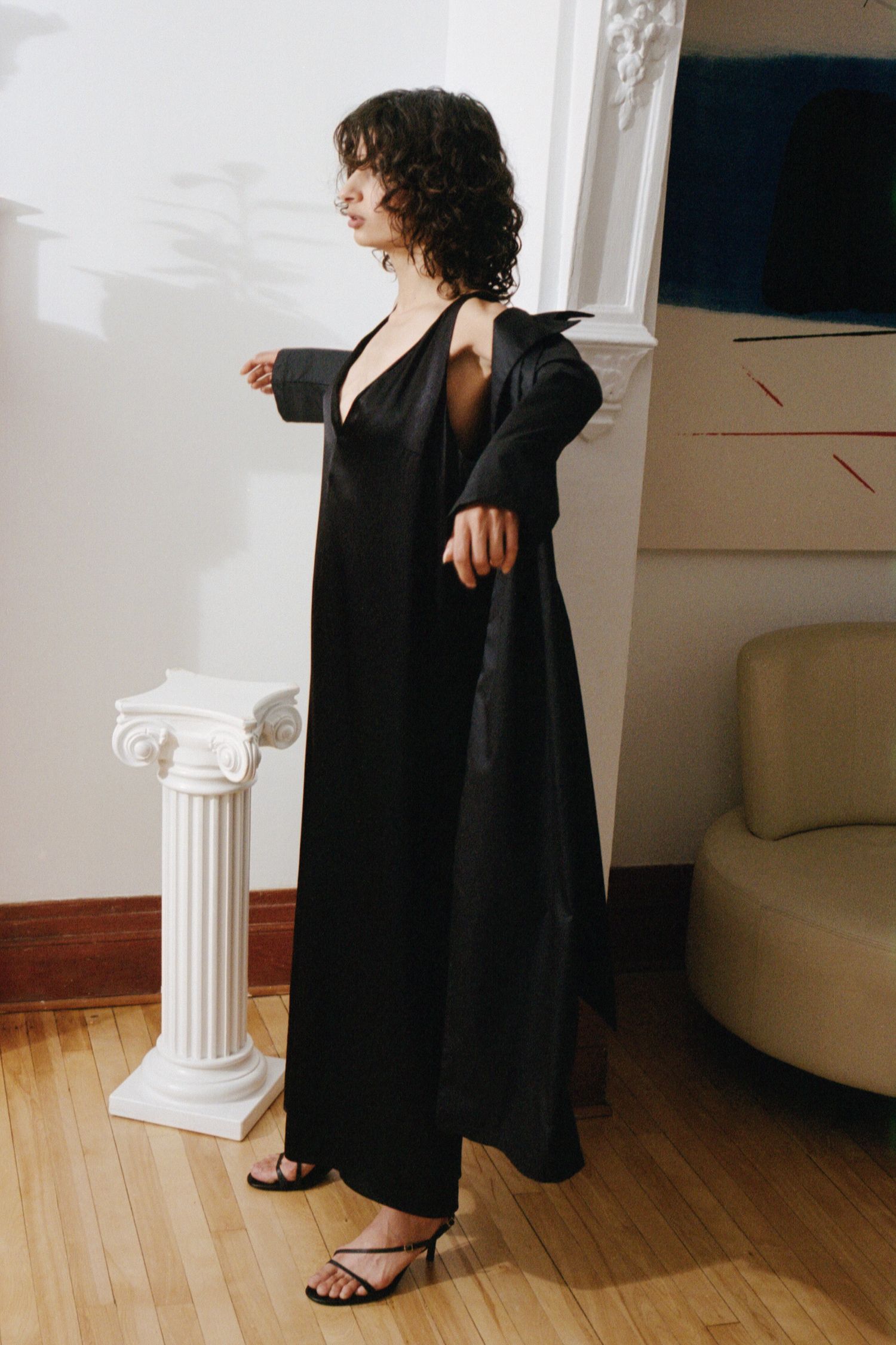 A model puts on a long black satin jacket overtop a long black minimalist dress from Waves of Hydra.