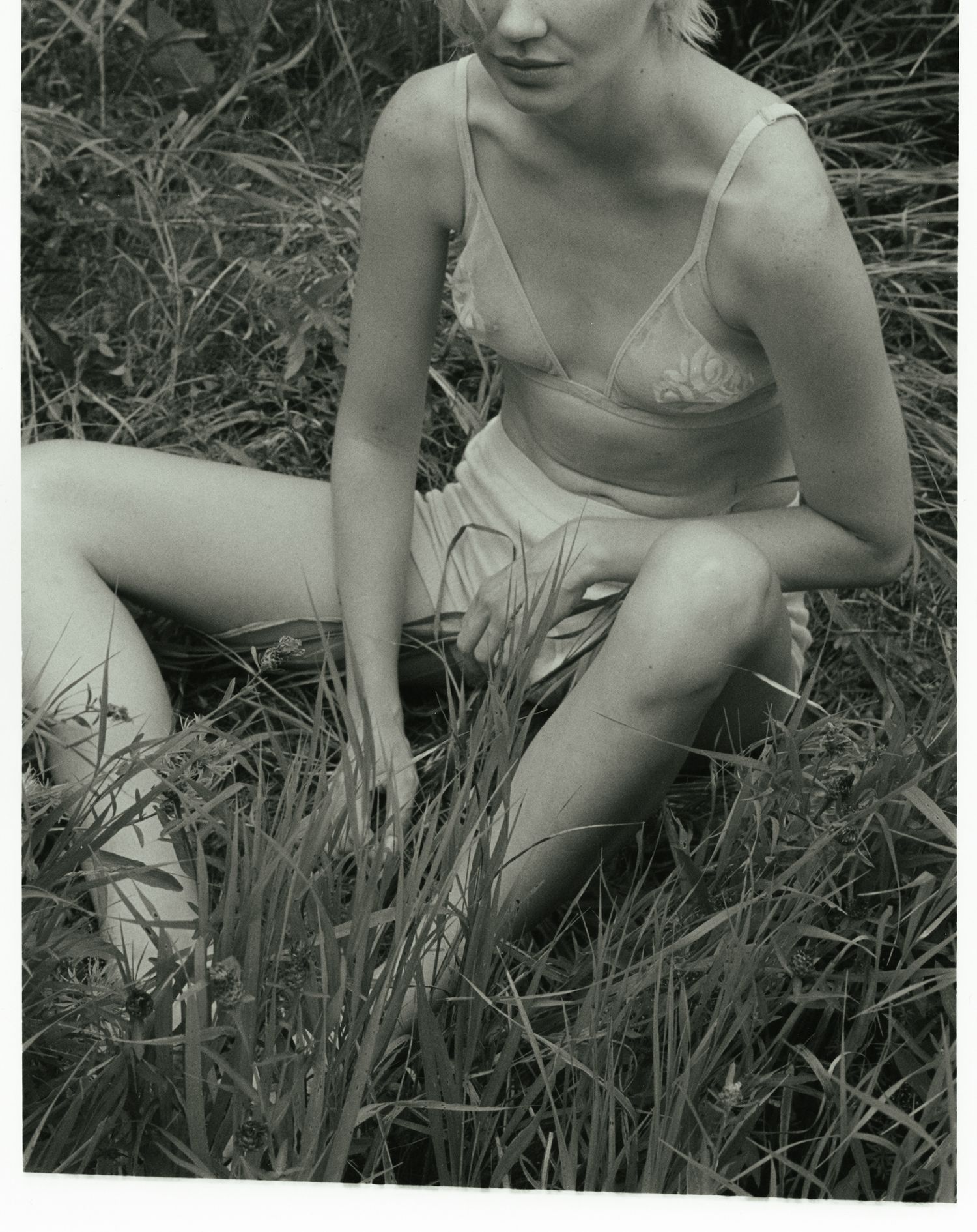 Black and white film portrait of a model sitting in a field of tall grass wearing a sheer vintage bra.