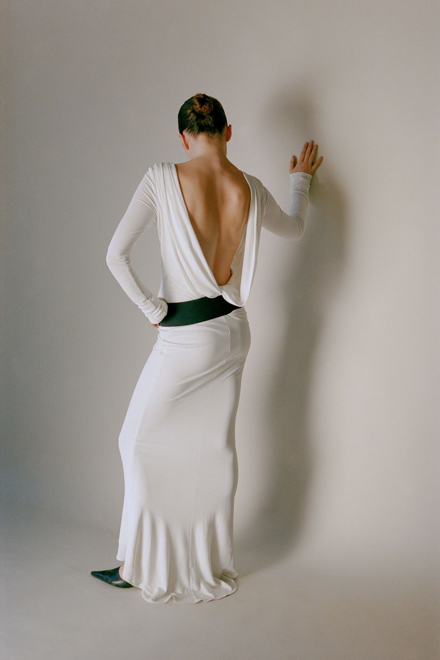 A model stands in a white studio with her back to the camera, wearing a long draped cream dress and large black belt.
