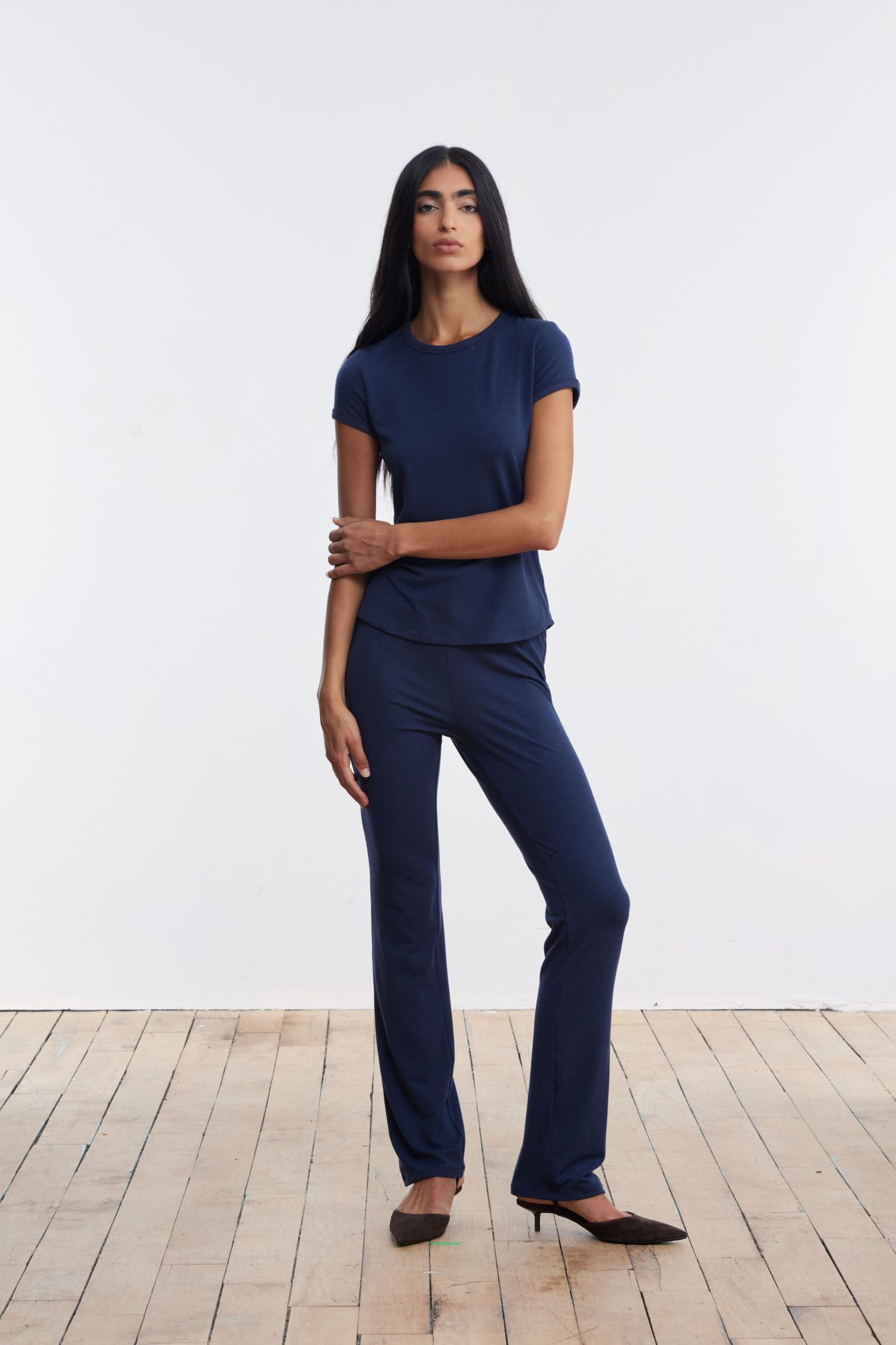 A model stands looking at the camera in a studio with wood flooring wearing a matching navy jersey tee and pant set.