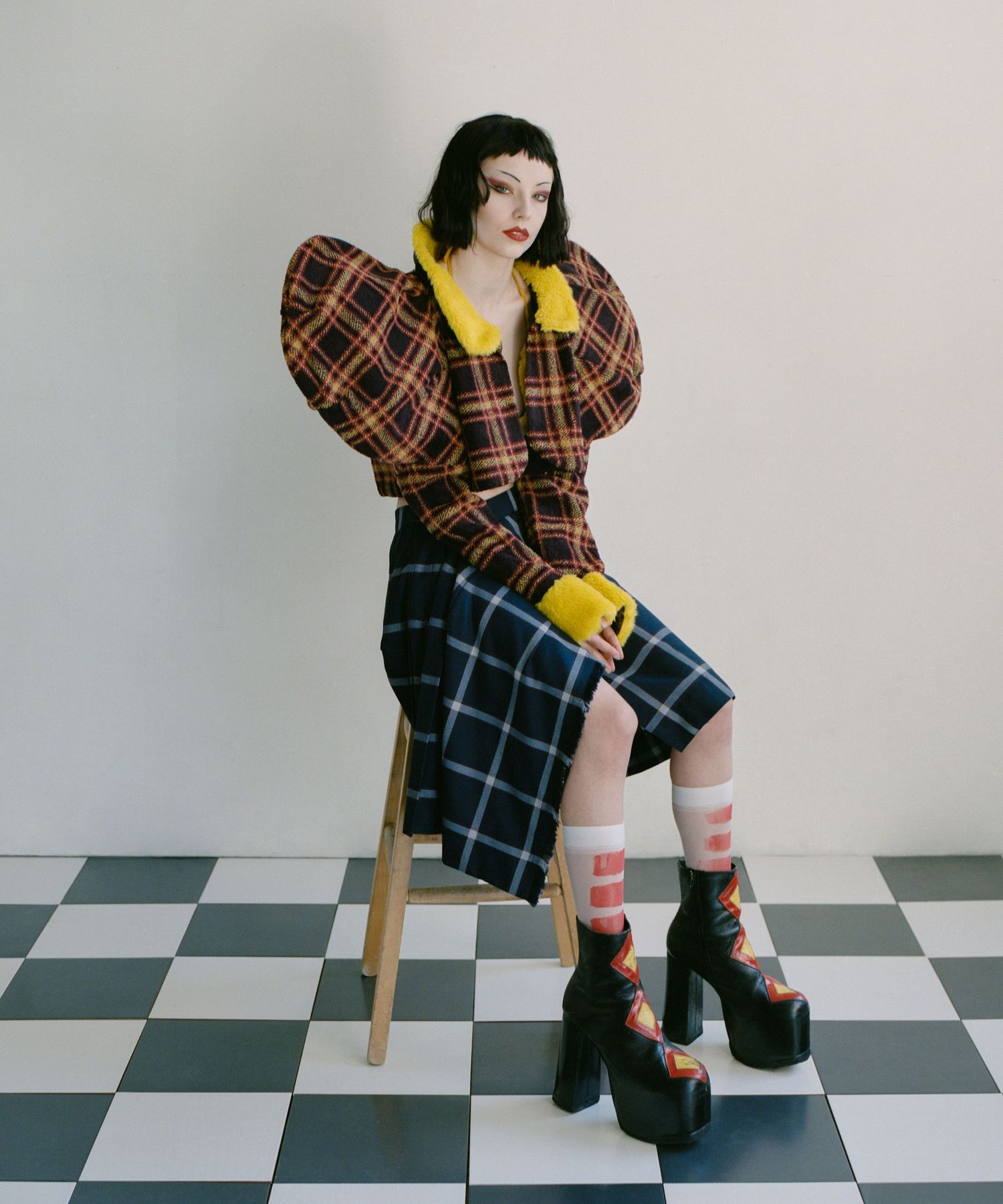 A model with a blunt short haircut sits on a stool wearing large platform shoes and a plaid outfit with an exaggerated silhouette.
