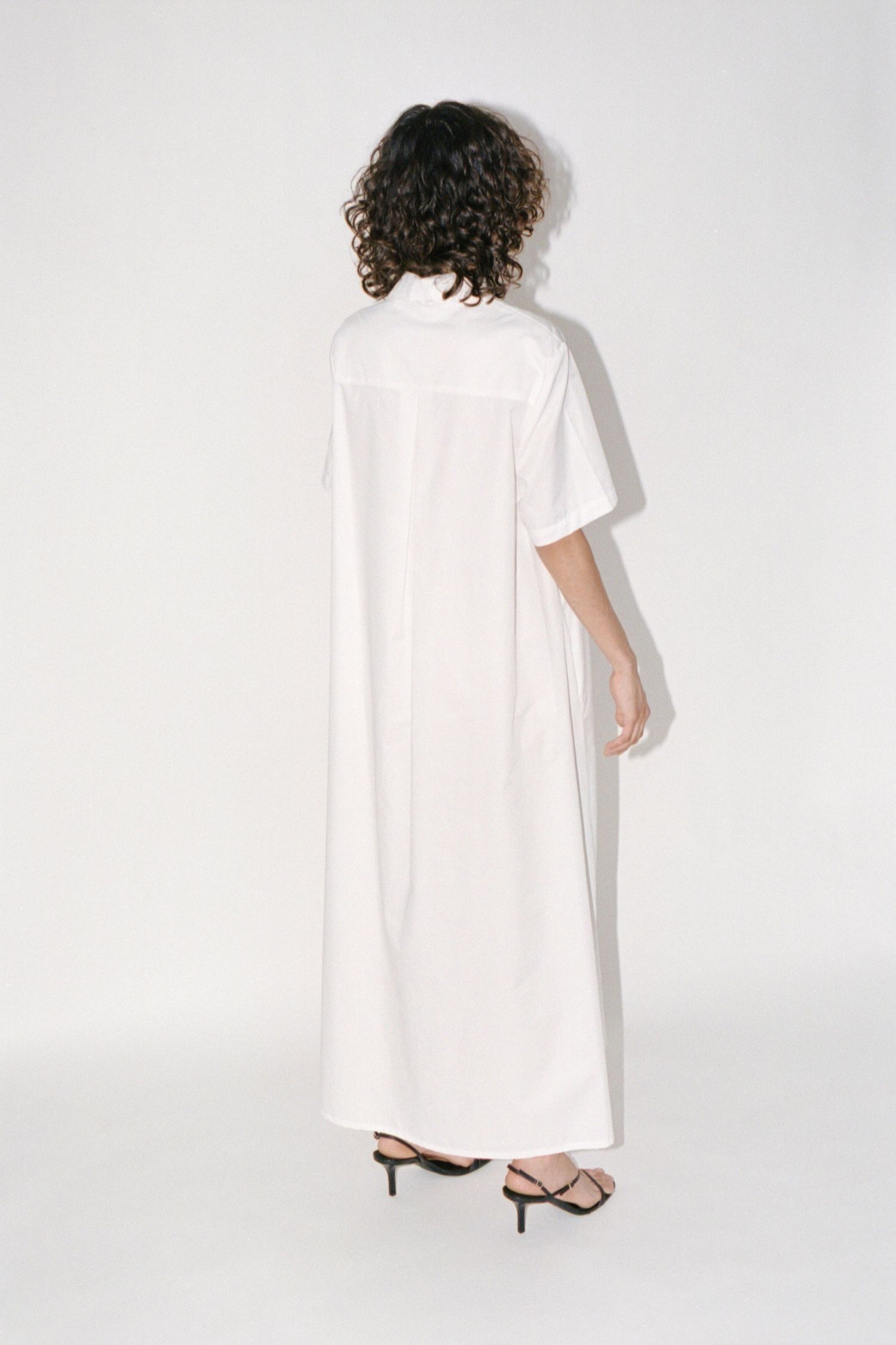 A wavy-haired brunette model wears a minimalistic long white cotton dress by Canadian brand Waves of Hydra.