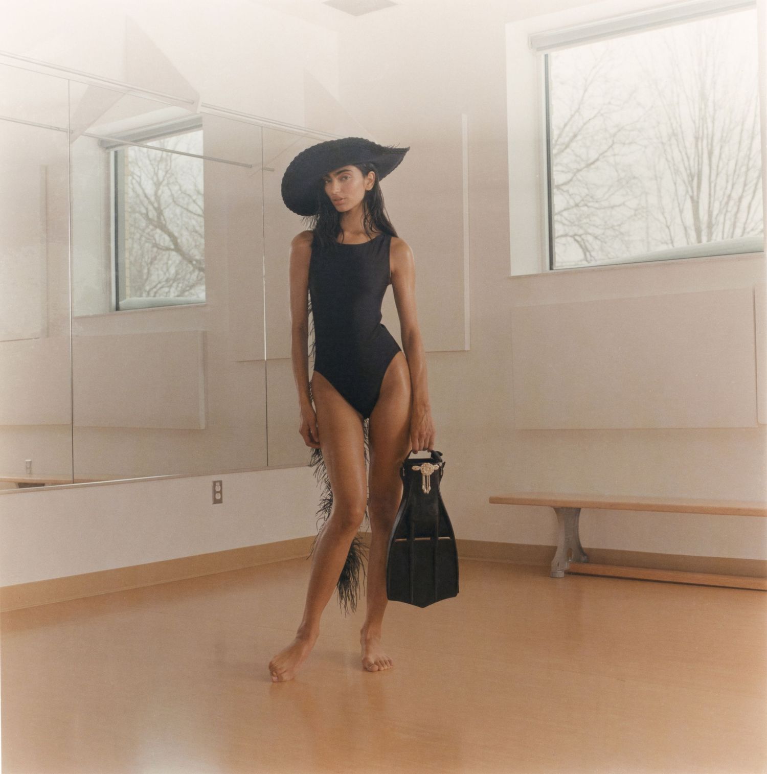 A model wearing a black one-piece swimsuit from Minnow Bathers holds a swimming flipper while posing in a mirrored dance studio.