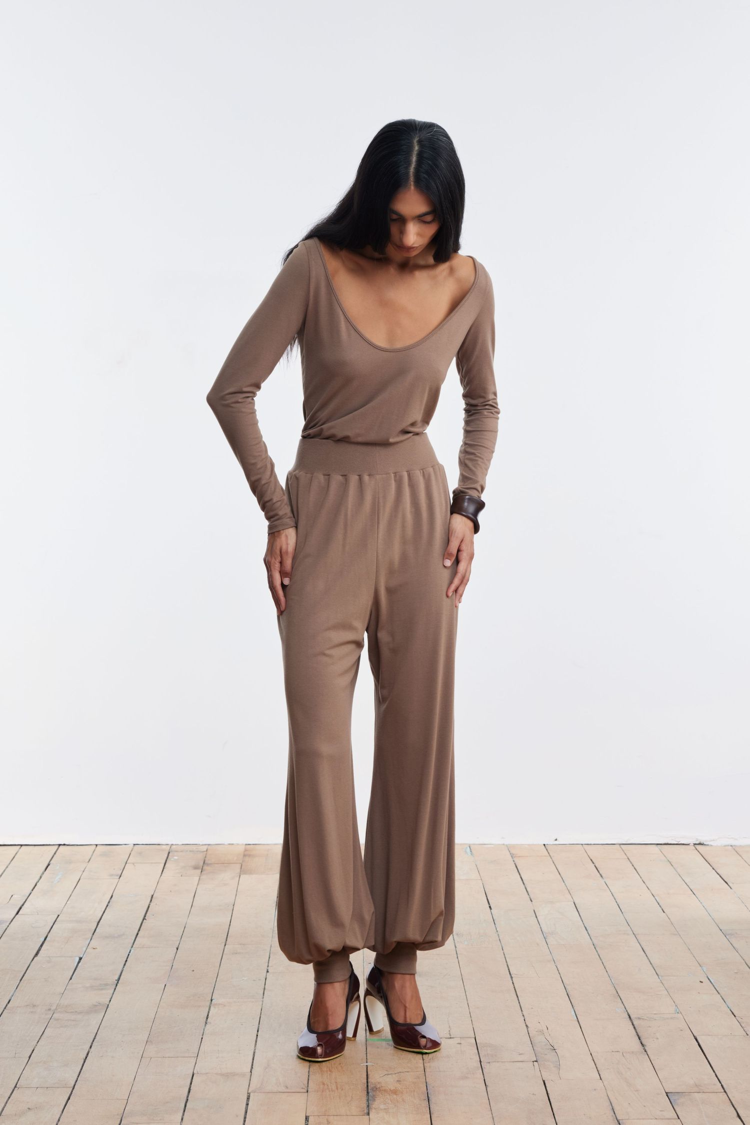 A model stands looking down in a studio wearing matching camel balloon pants and long sleeve scoop shirt.