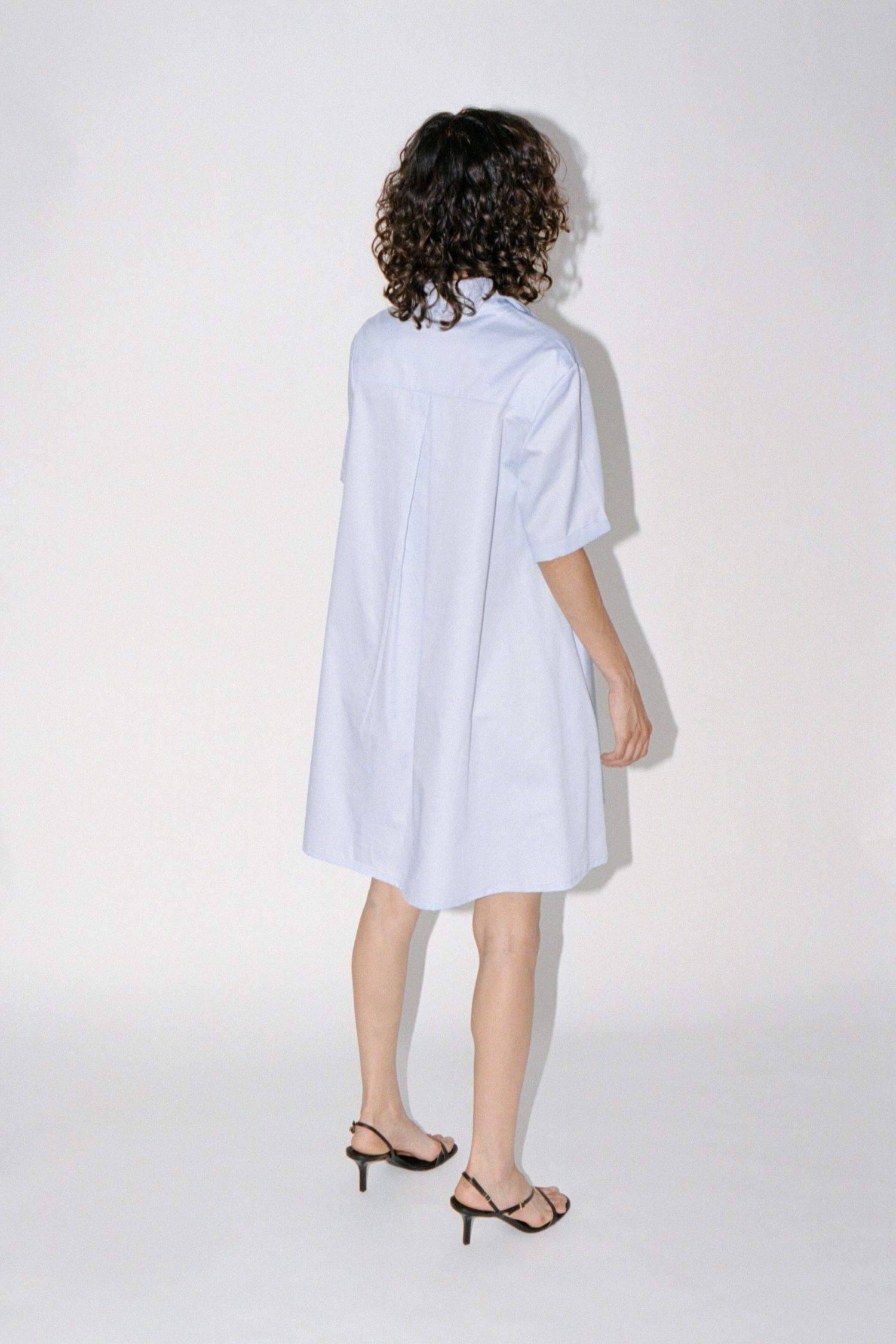 A brunette model faces backward wearing a baby blue short cotton dress by Montreal brand Waves of Hydra.