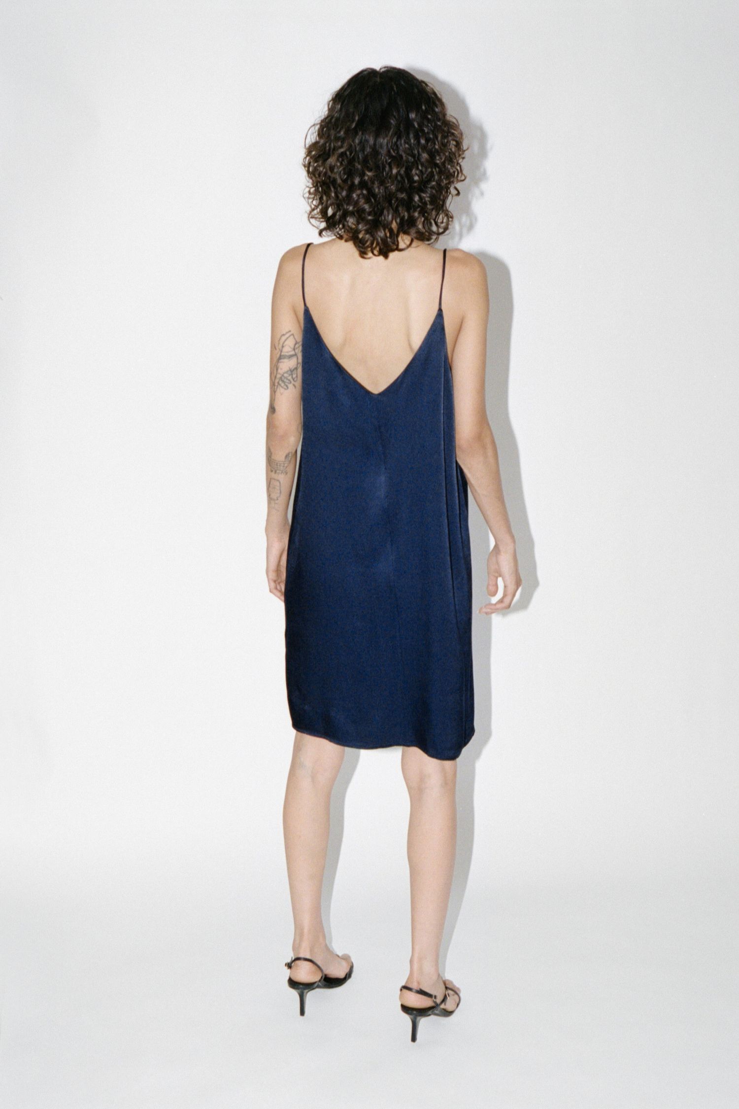 A wavy-haired brunette model wears a navy satin slip dress with a low back by Waves of Hydra.