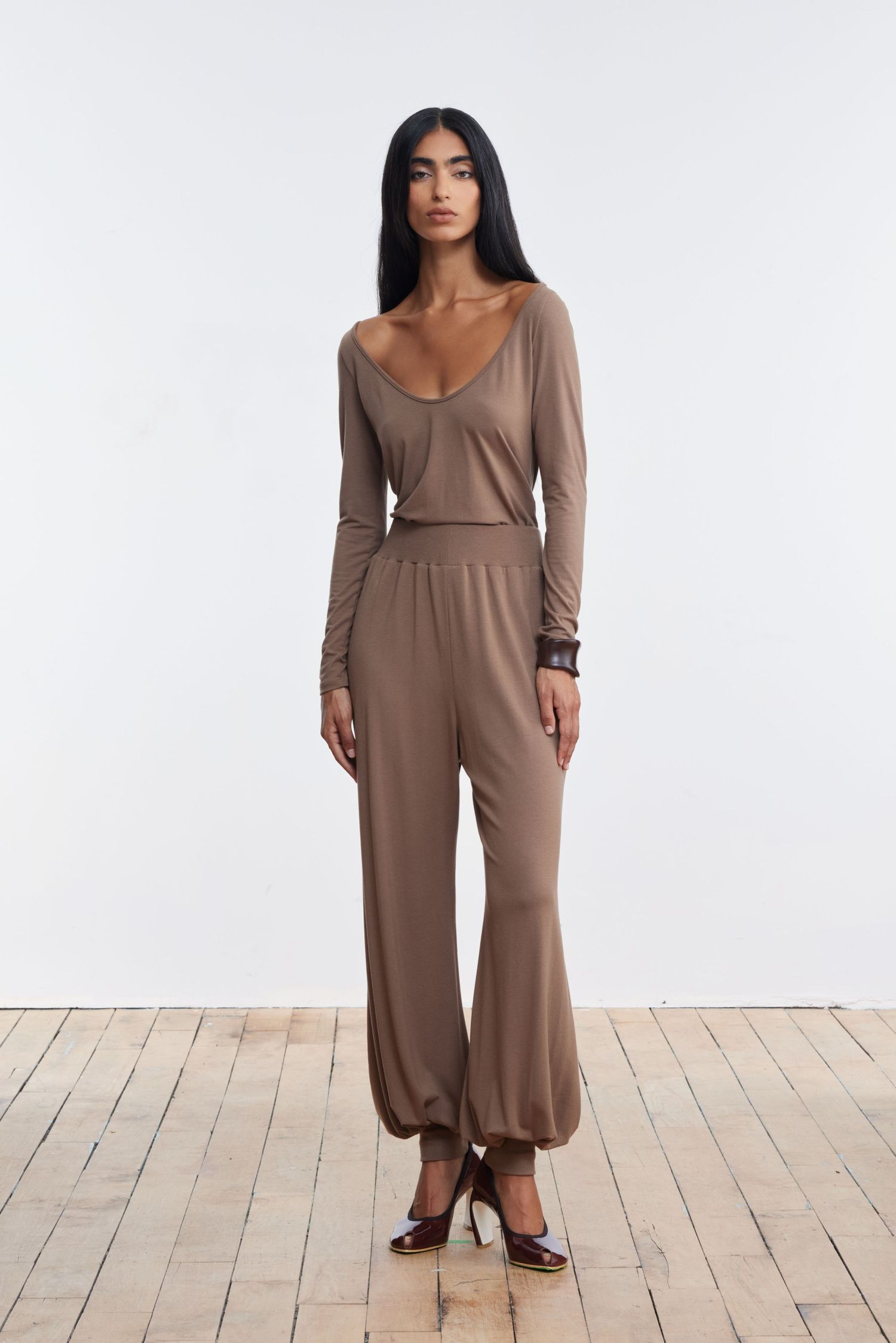 A model stands looking at the camera in a studio with wood flooring wearing matching tan balloon pants and a long sleeve scoop shirt.