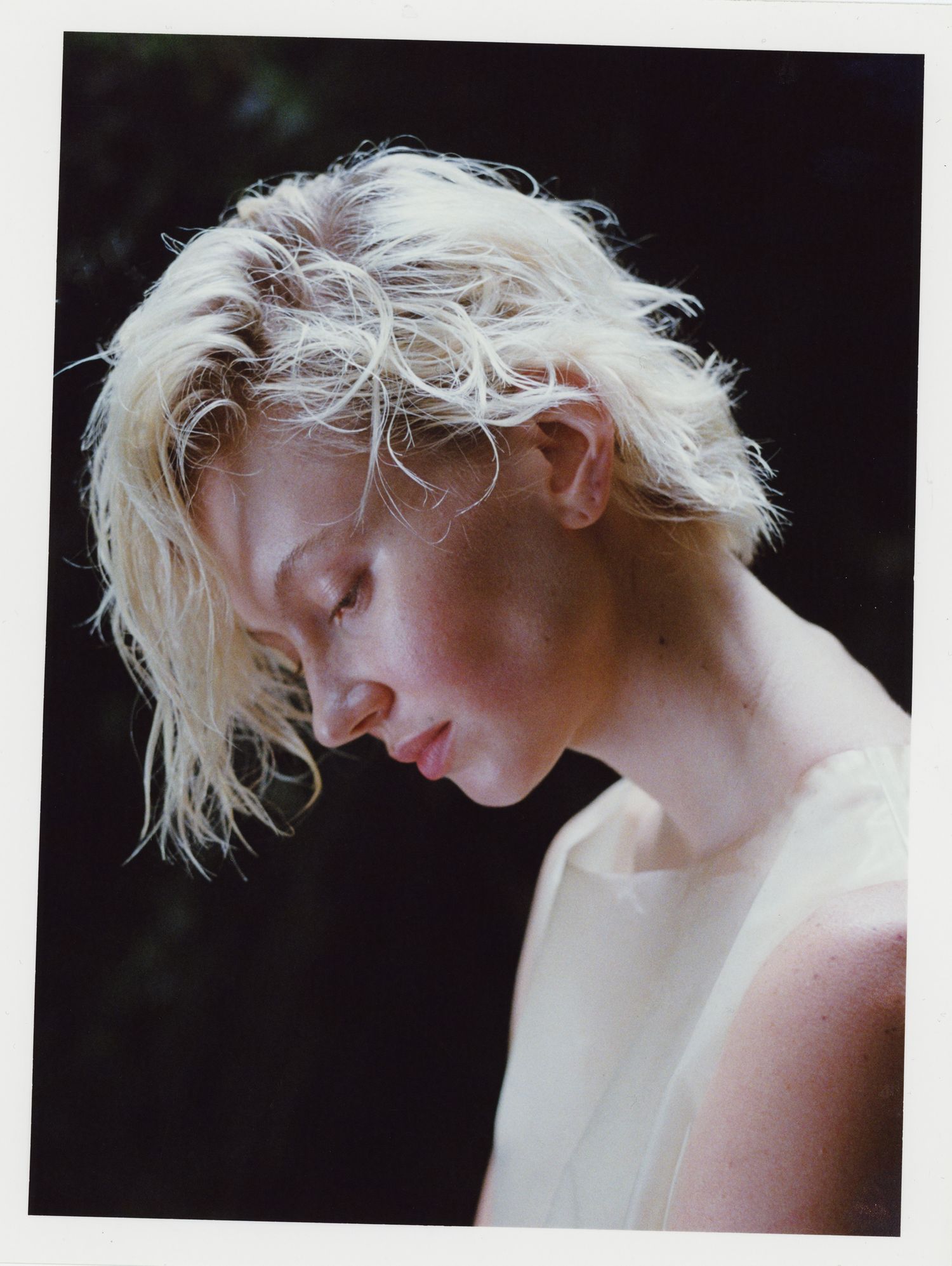 A traditional color darkroom print of a platinum blonde model wearing a translucent latex dress looking down, illuminated by sunlight.