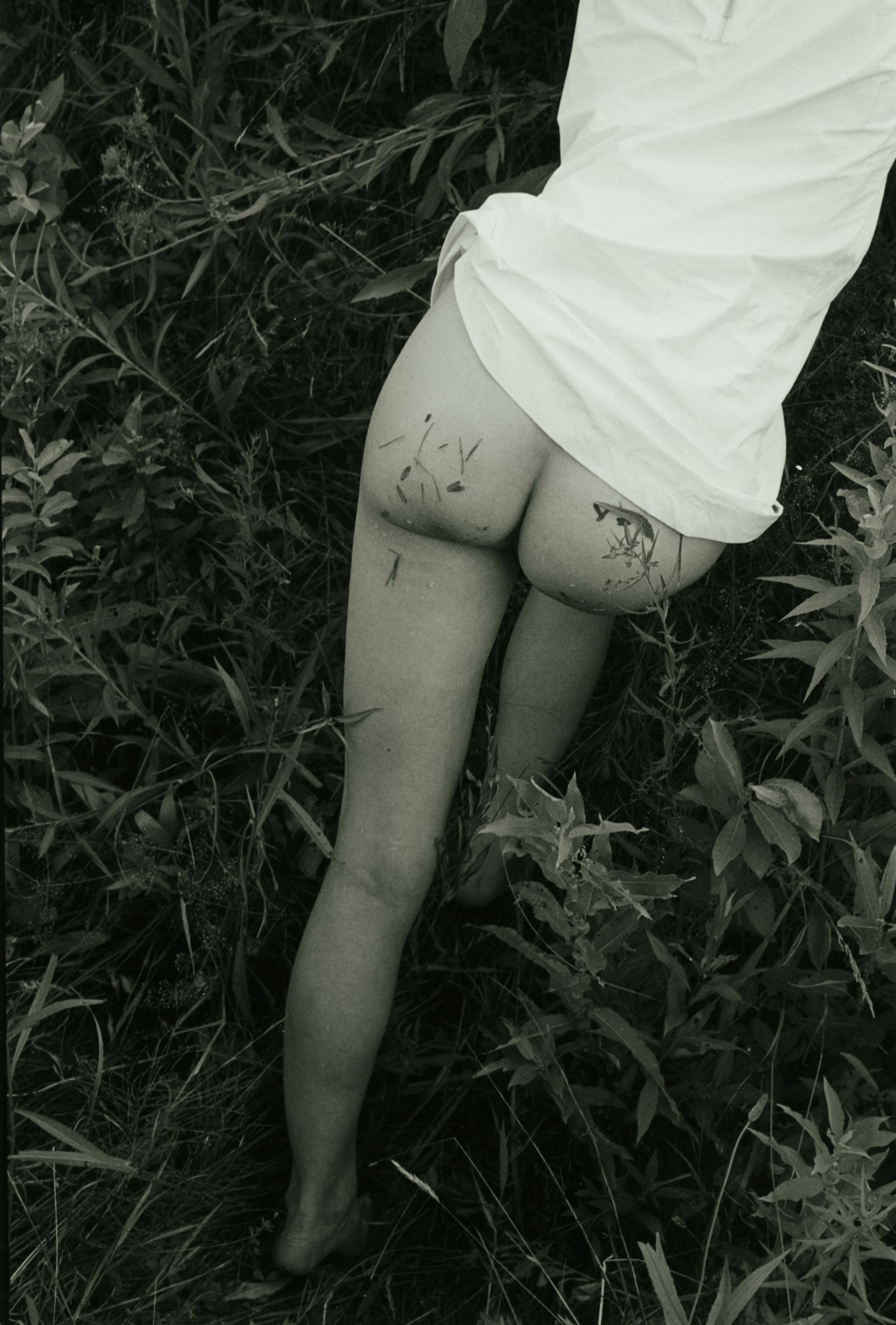 Black and white film portrait of a model in a plastic dress standing in tall grass with her legs and bottom covered in grass.
