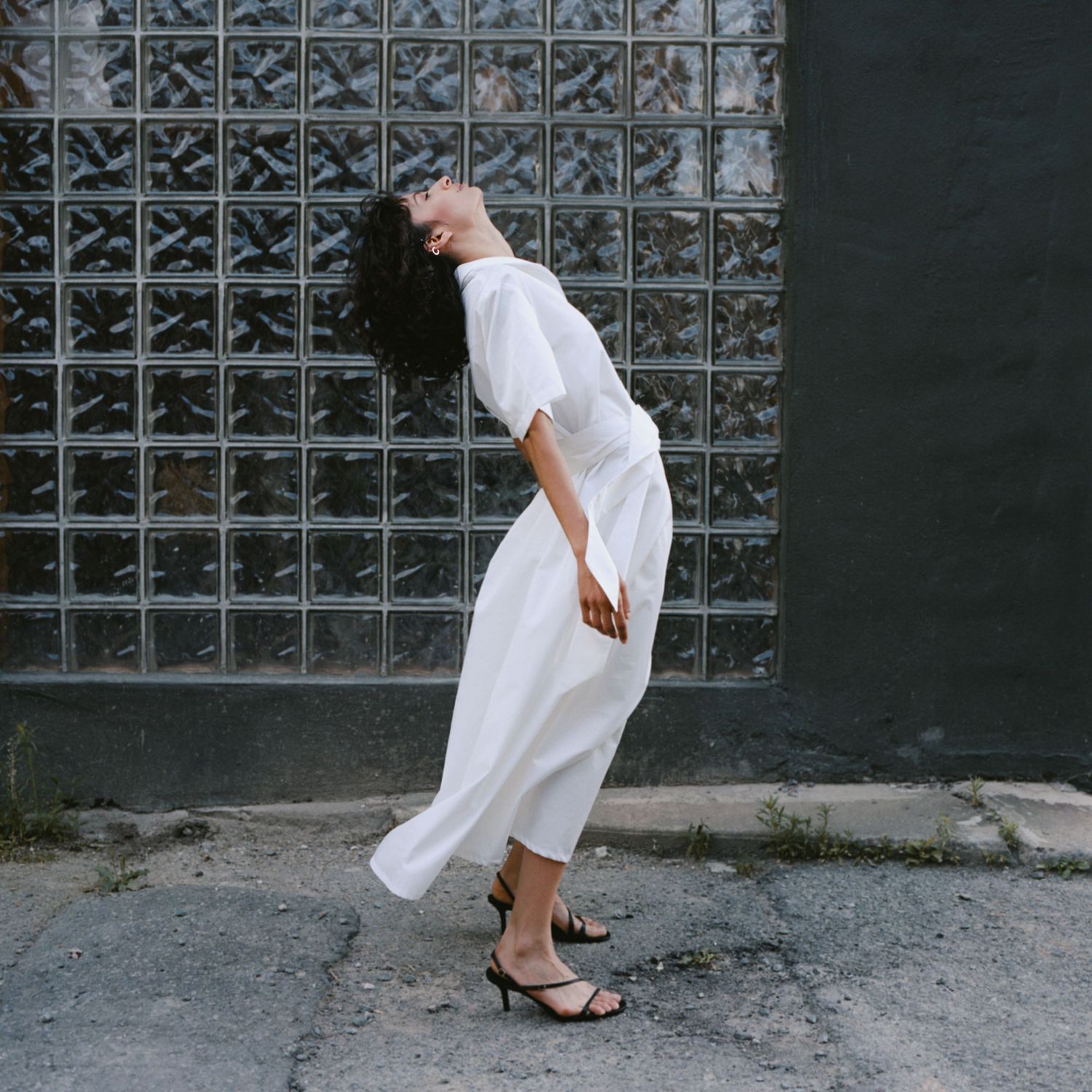 A model folds bends backward while wearing a long white crisp cotton dress and strappy heels in front of a green wall with glass bricks.