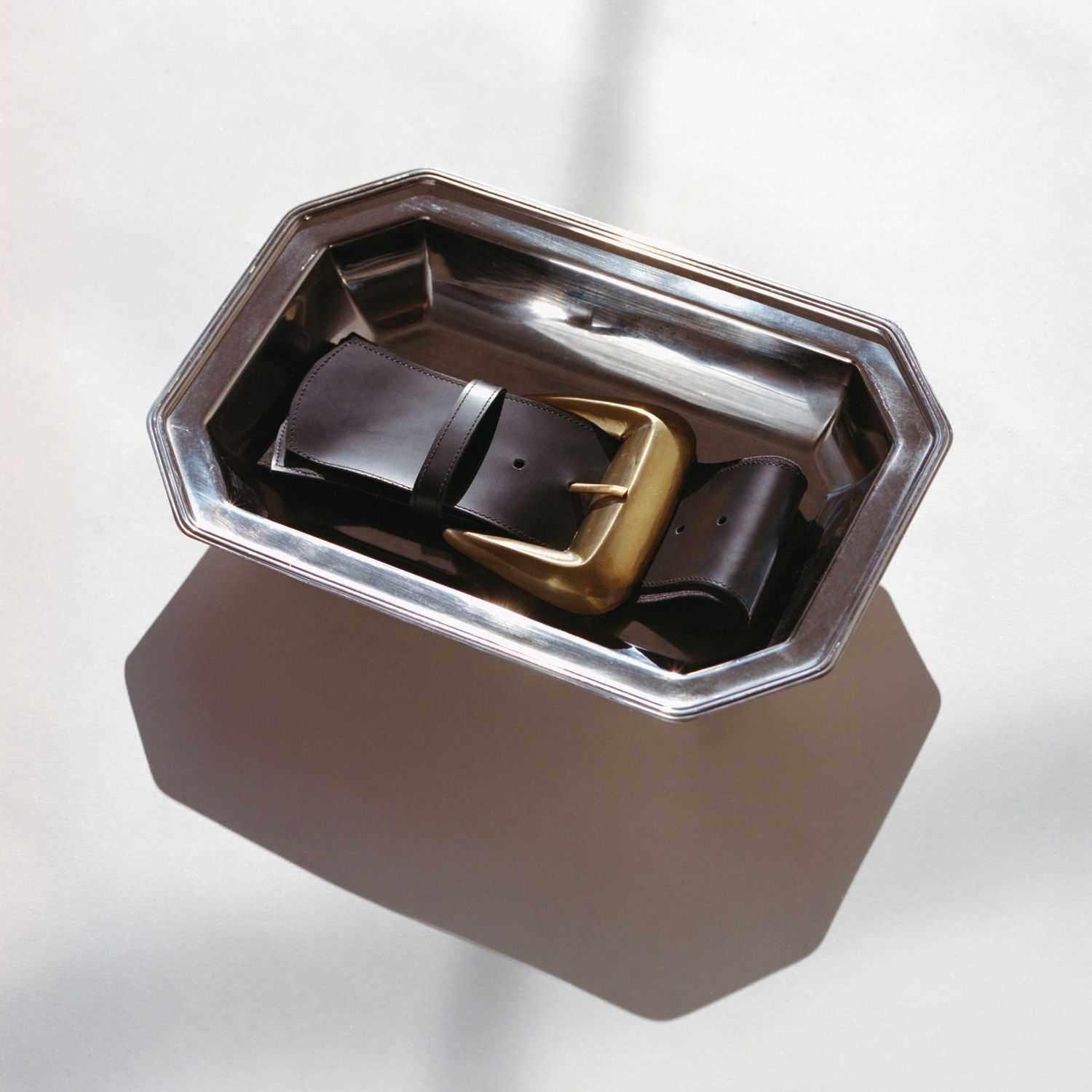 A black leather belt with an oversized brass buckle by Atelier Jéa sits on a sterling silver tray in the sunlight.