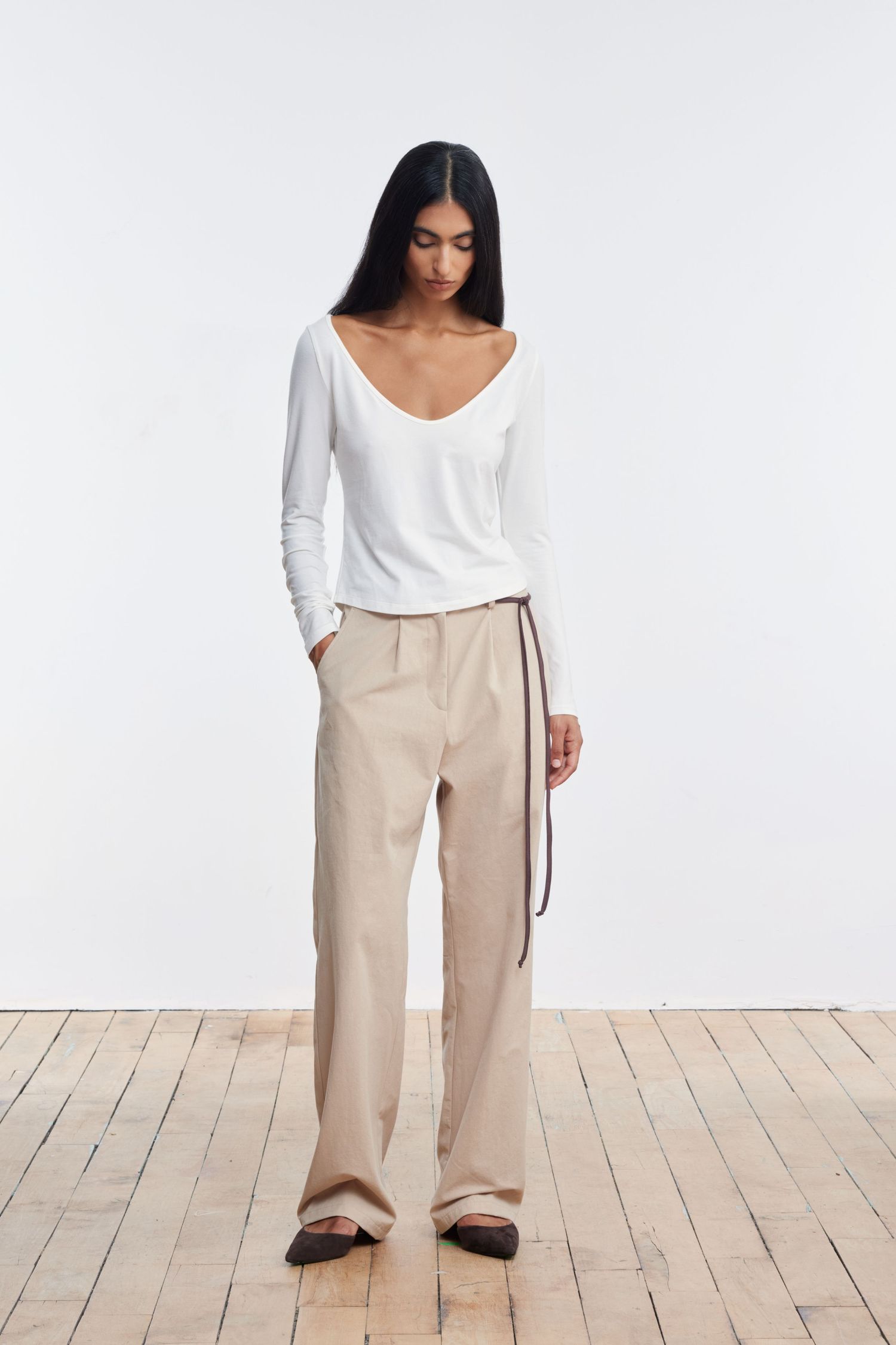 A model stands, looking down, in a wood-floored studio wearing khaki pleated cotton trousers and a white cotton scoop tee.