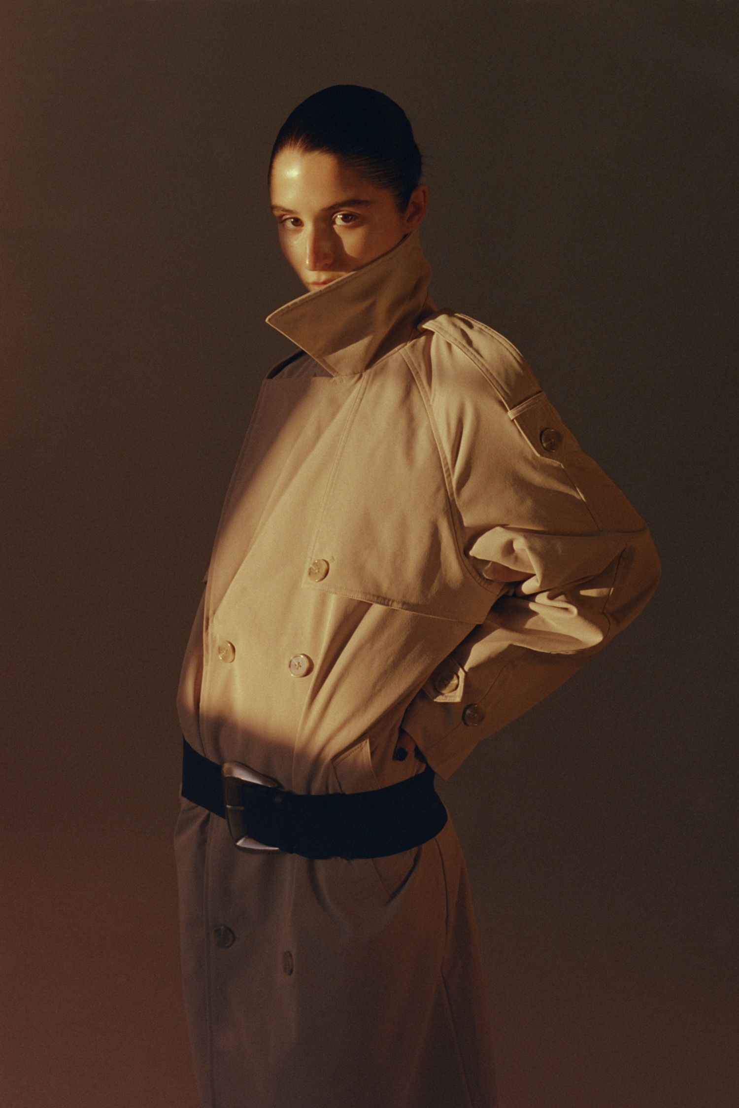 A model poses in a camel trench coat in studio with natural golden hour illuminating her face.