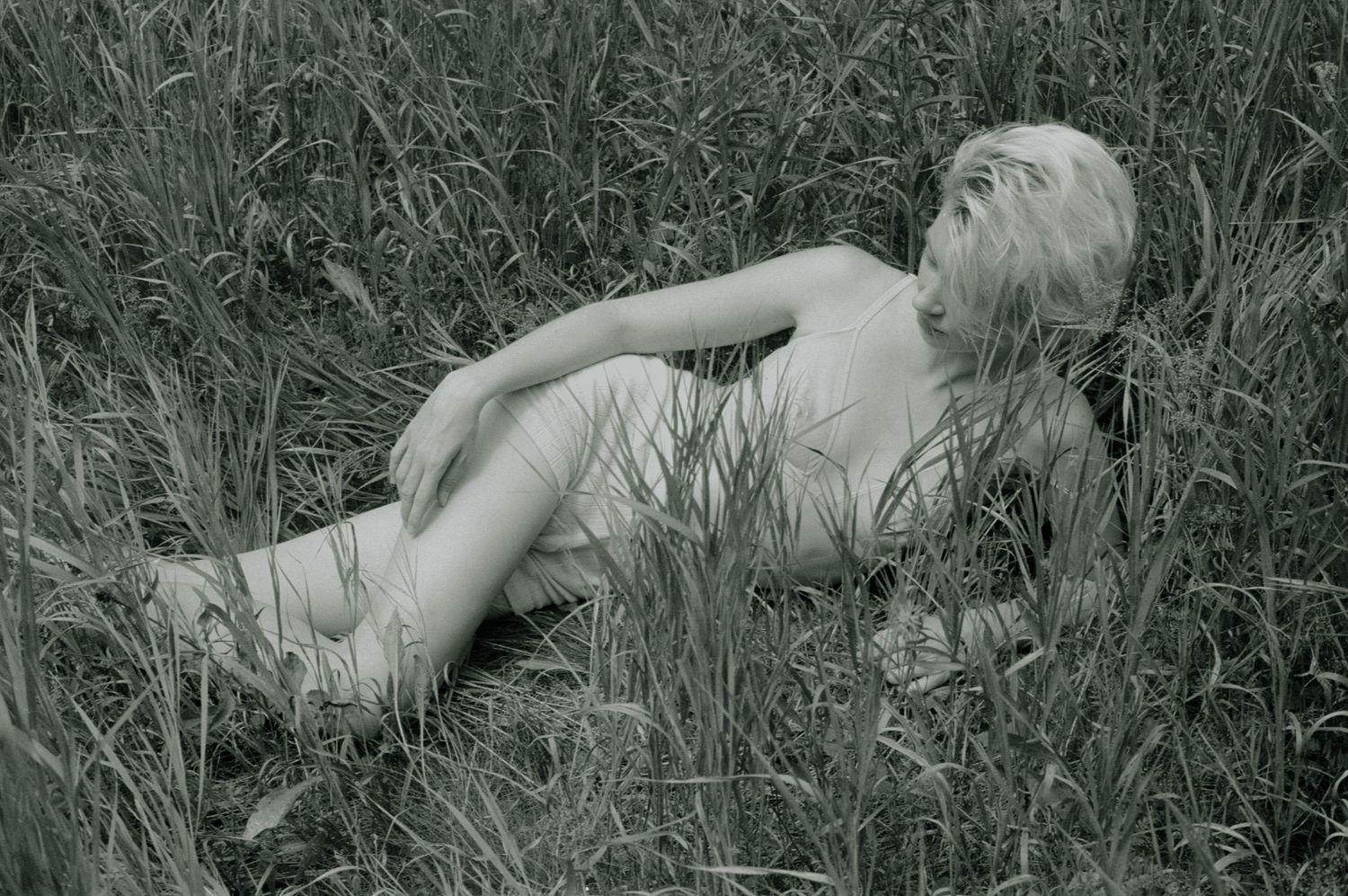 Black and white film portrait of a model lying on her side, partially obscured by tall grass.