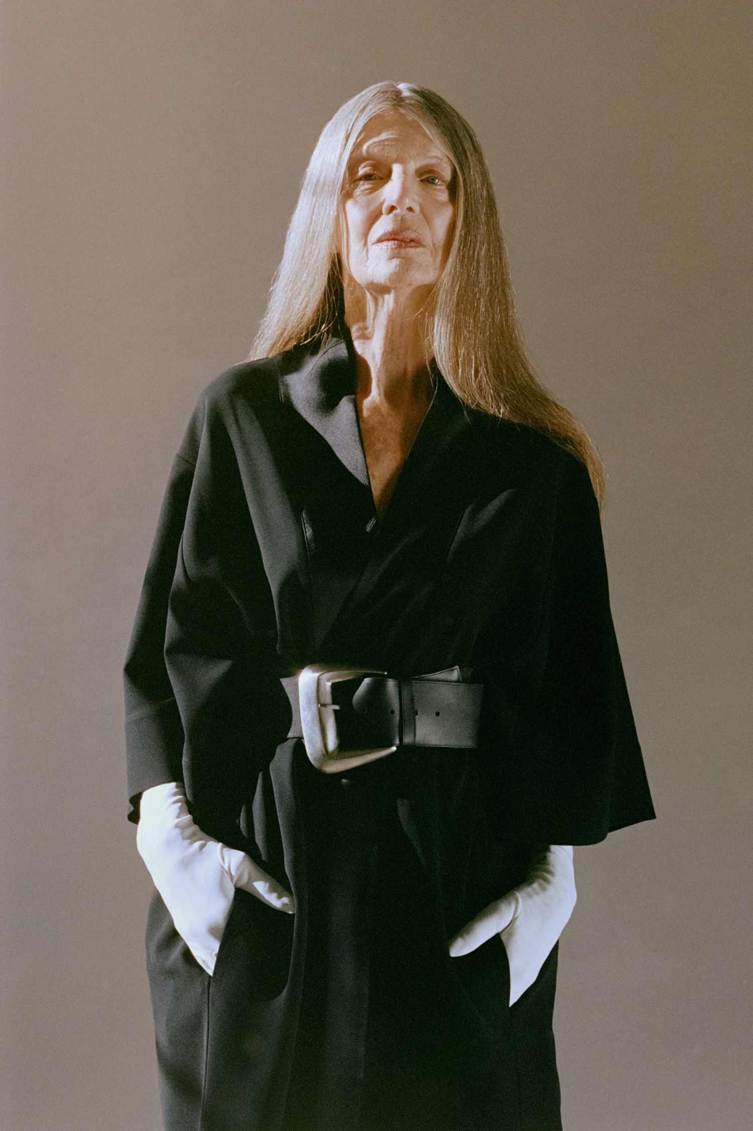 A model with long silver hair poses in studio wearing a black dress, white gloves, and oversized silver belt.