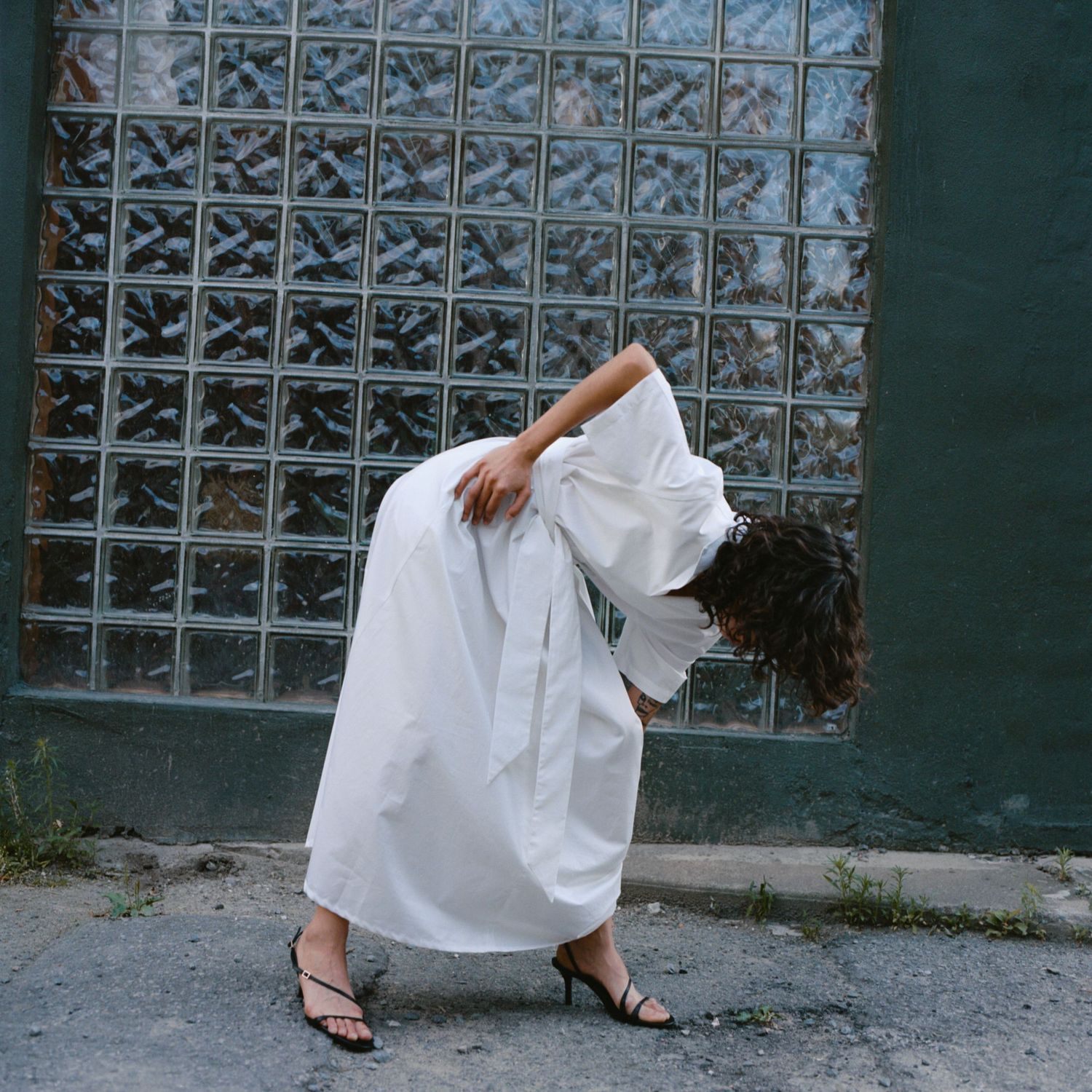 A model folds forward while wearing a long white crisp cotton dress and strappy heels.