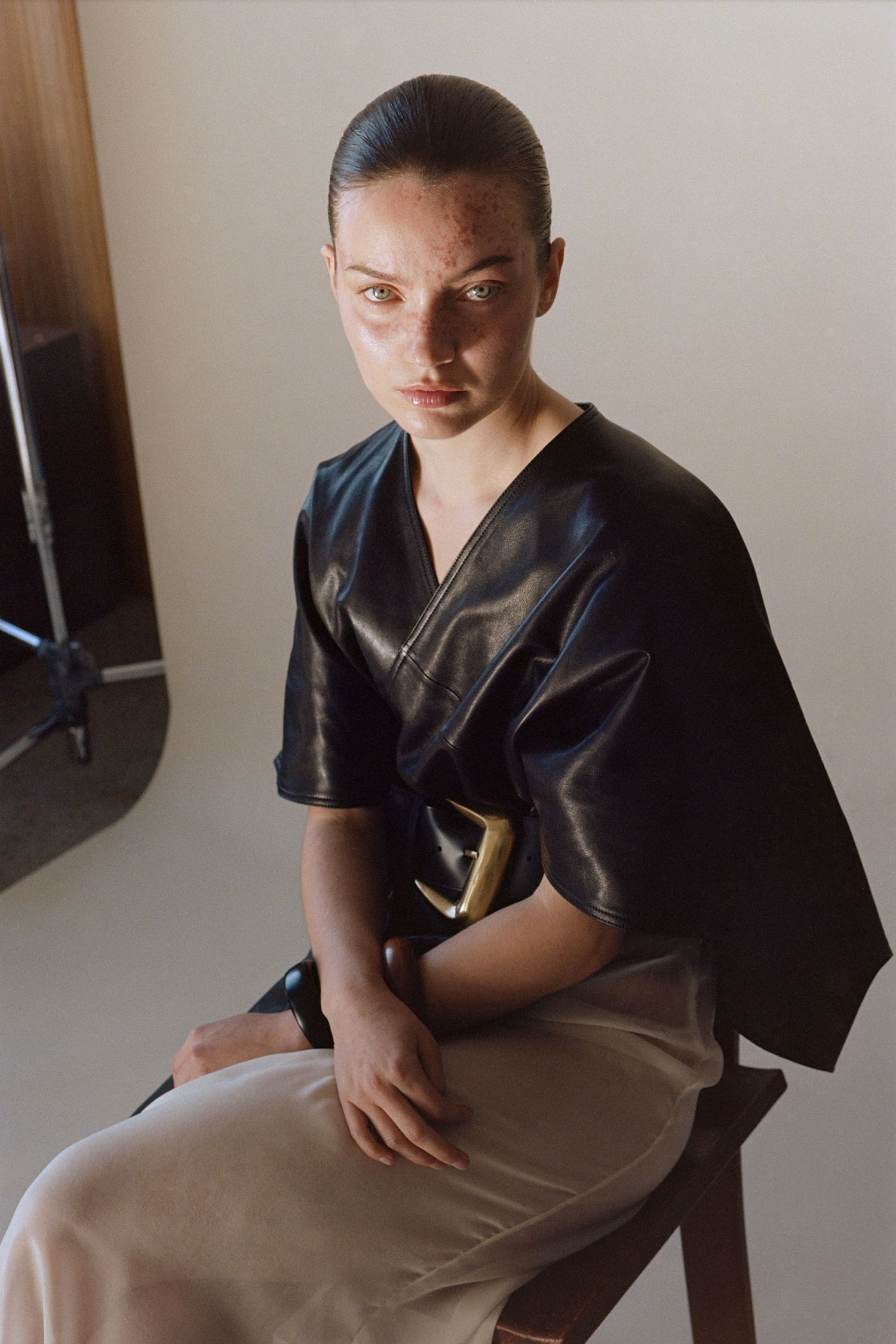 A model with freckles wears a leather kimono, cream silk slip skirt, and Atelier Jéa belt while sitting in a photo studio.