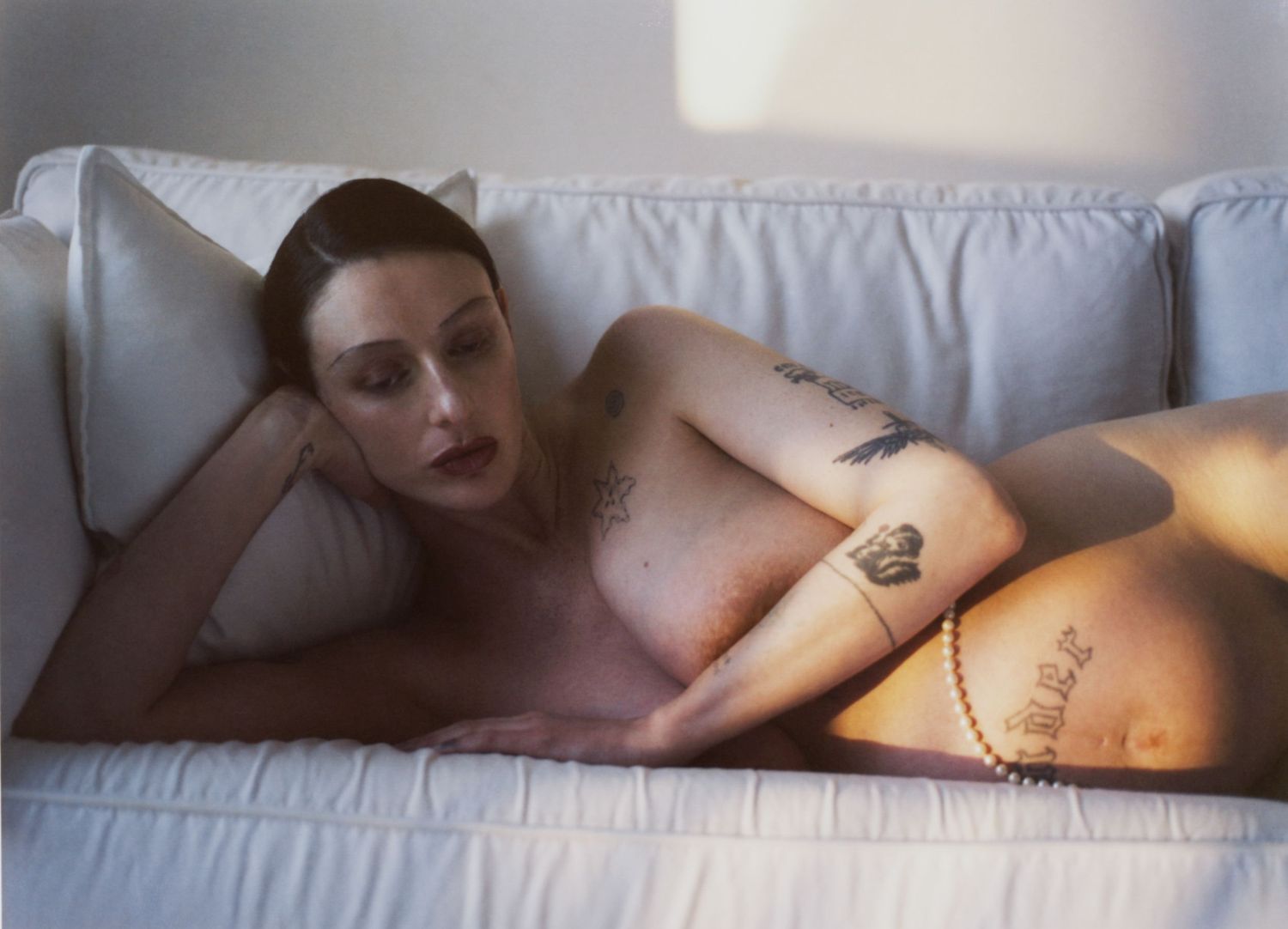 A pregnant woman lies nude on a white sofa while illuminated by golden sunlight.